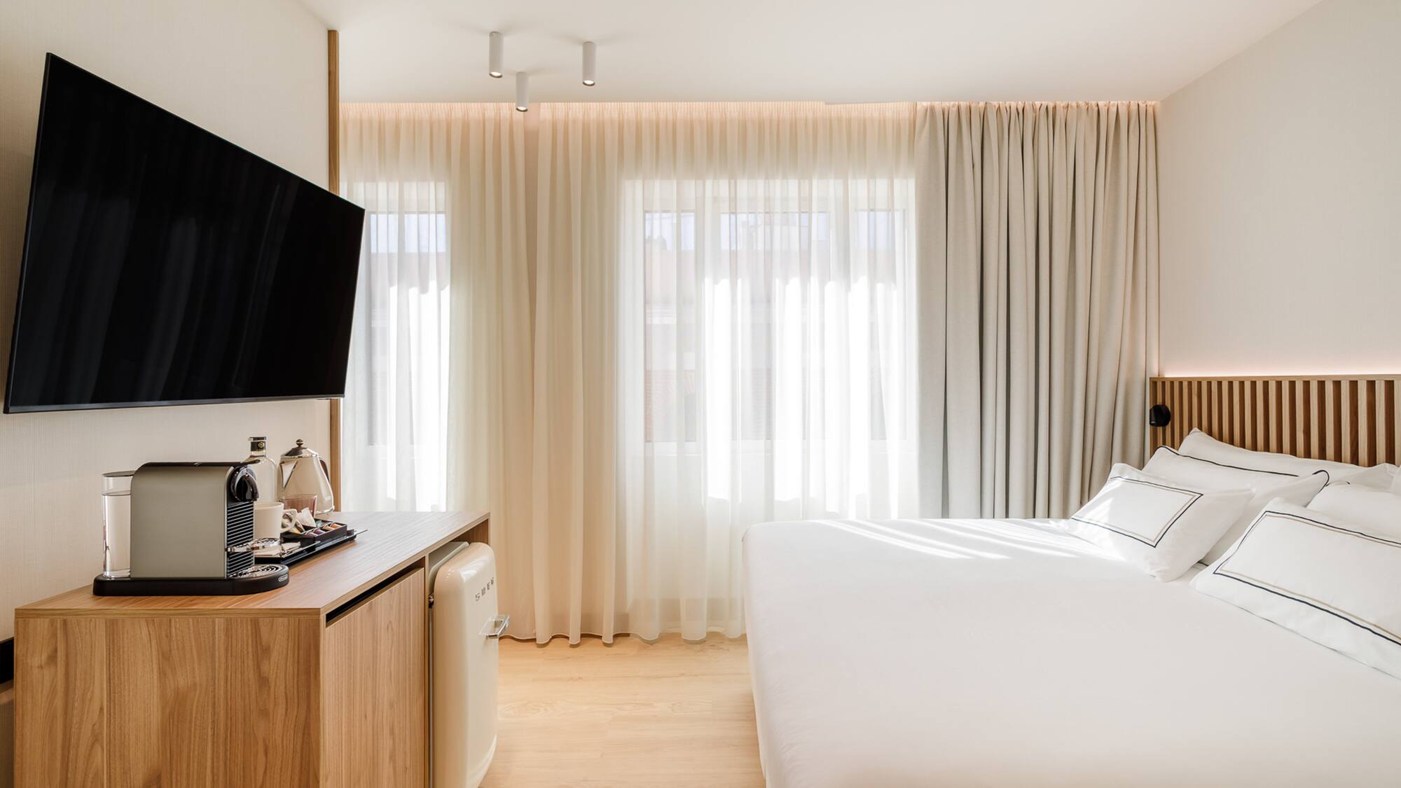 Modern hotel bedroom with TV, coffee machine, mini fridge, and white bed.