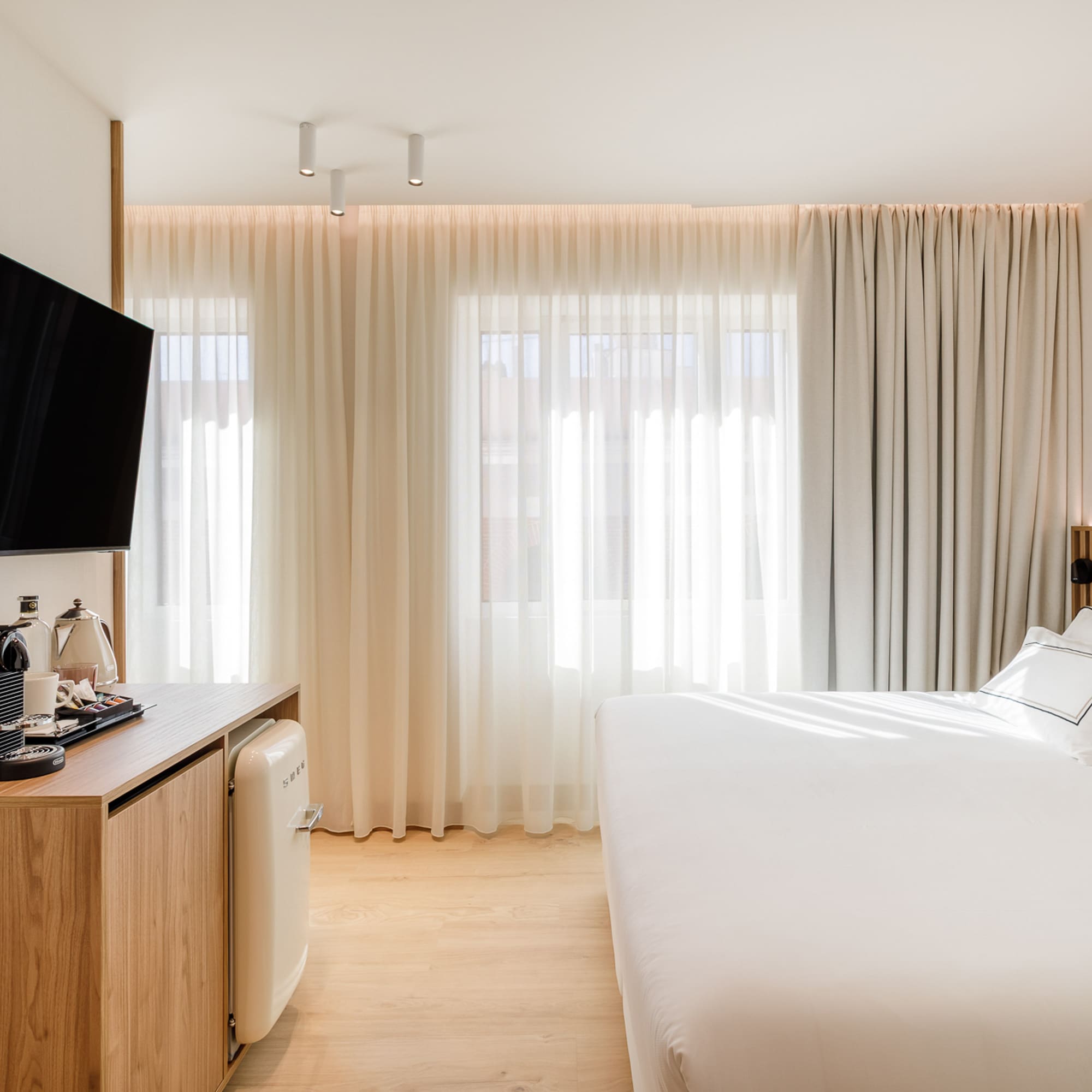 Modern hotel bedroom with TV, coffee machine, mini fridge, and white bed.