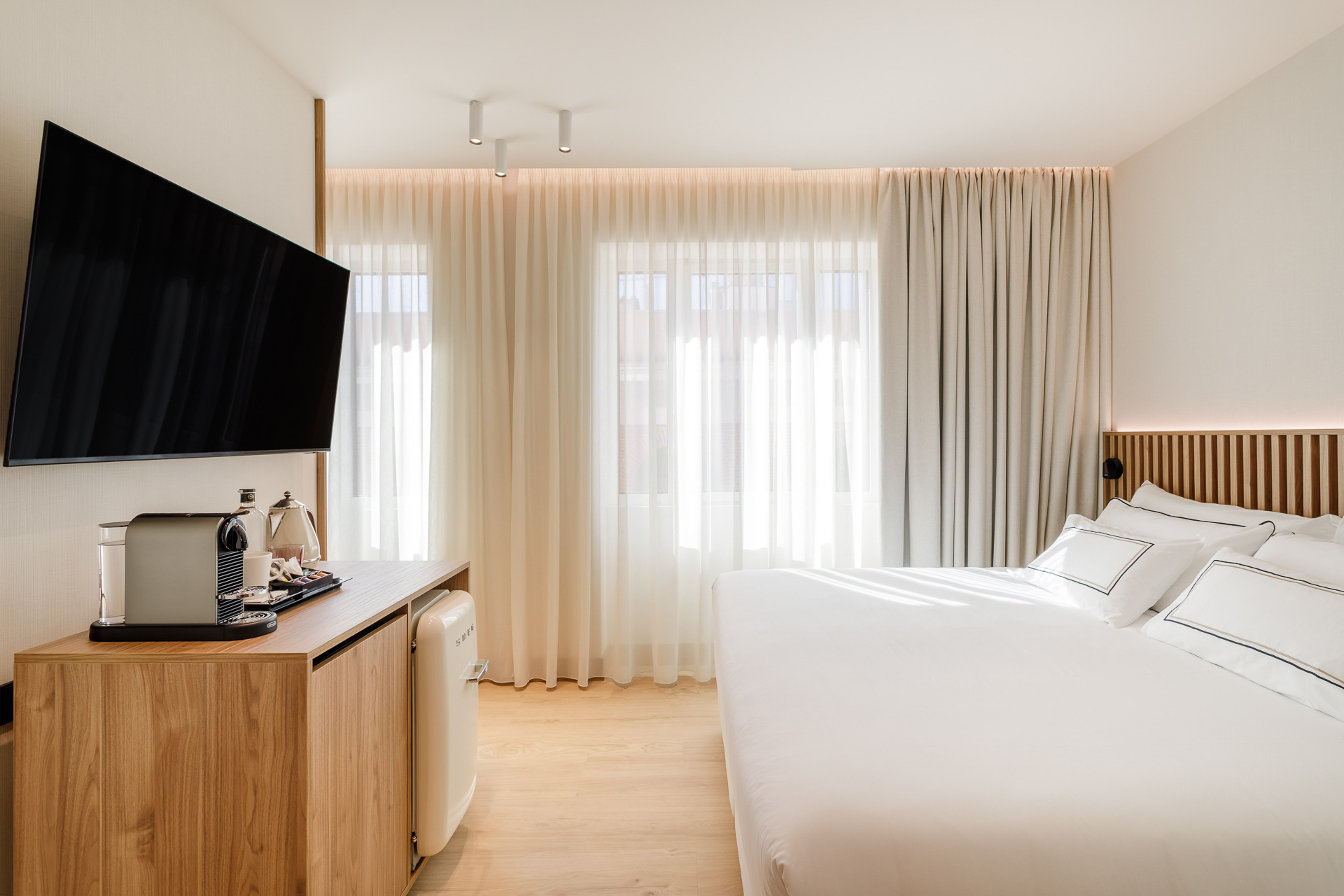 Modern hotel bedroom with TV, coffee machine, mini fridge, and white bed.
