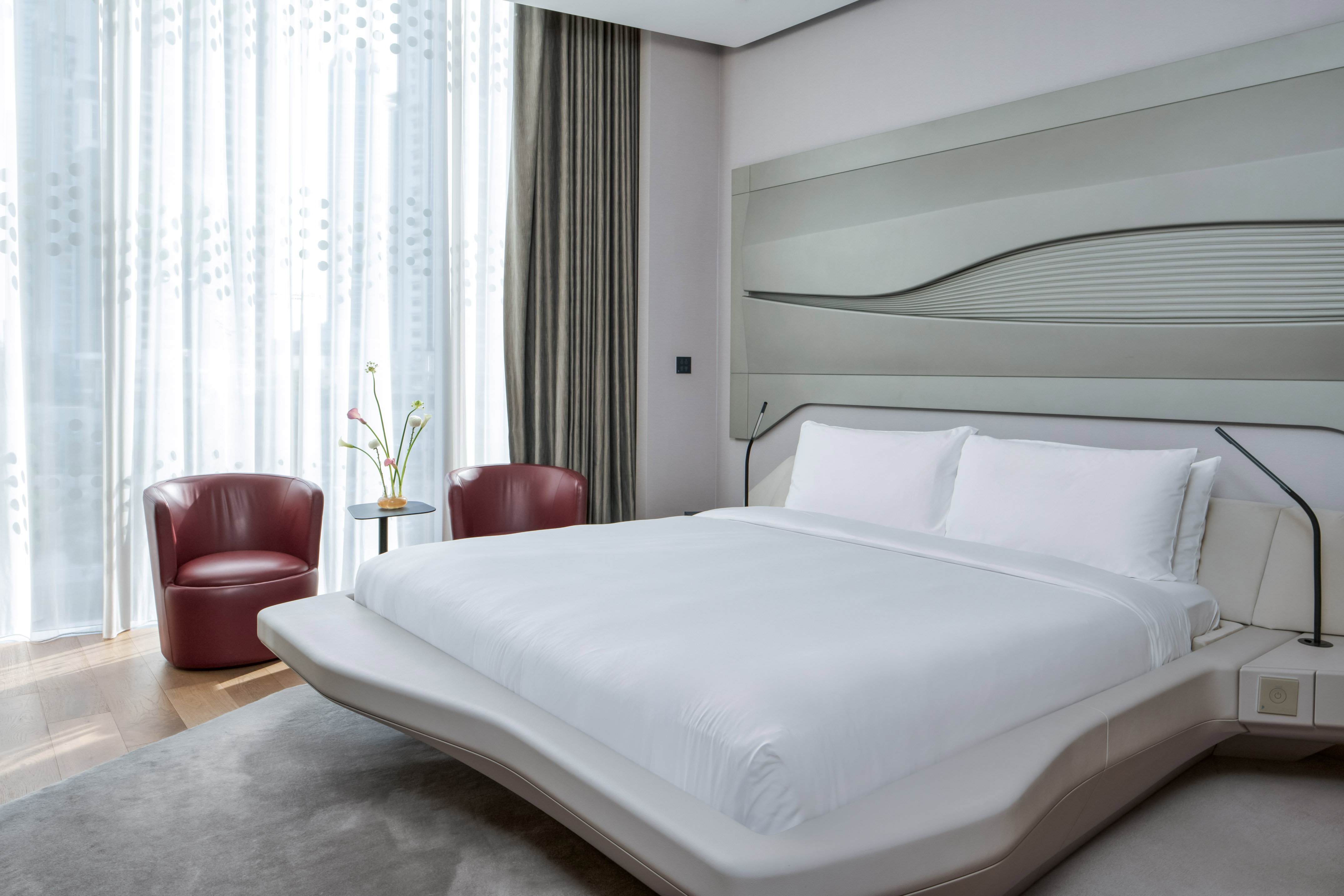 Modern hotel bedroom with white bed, wavy headboard, and red armchairs.