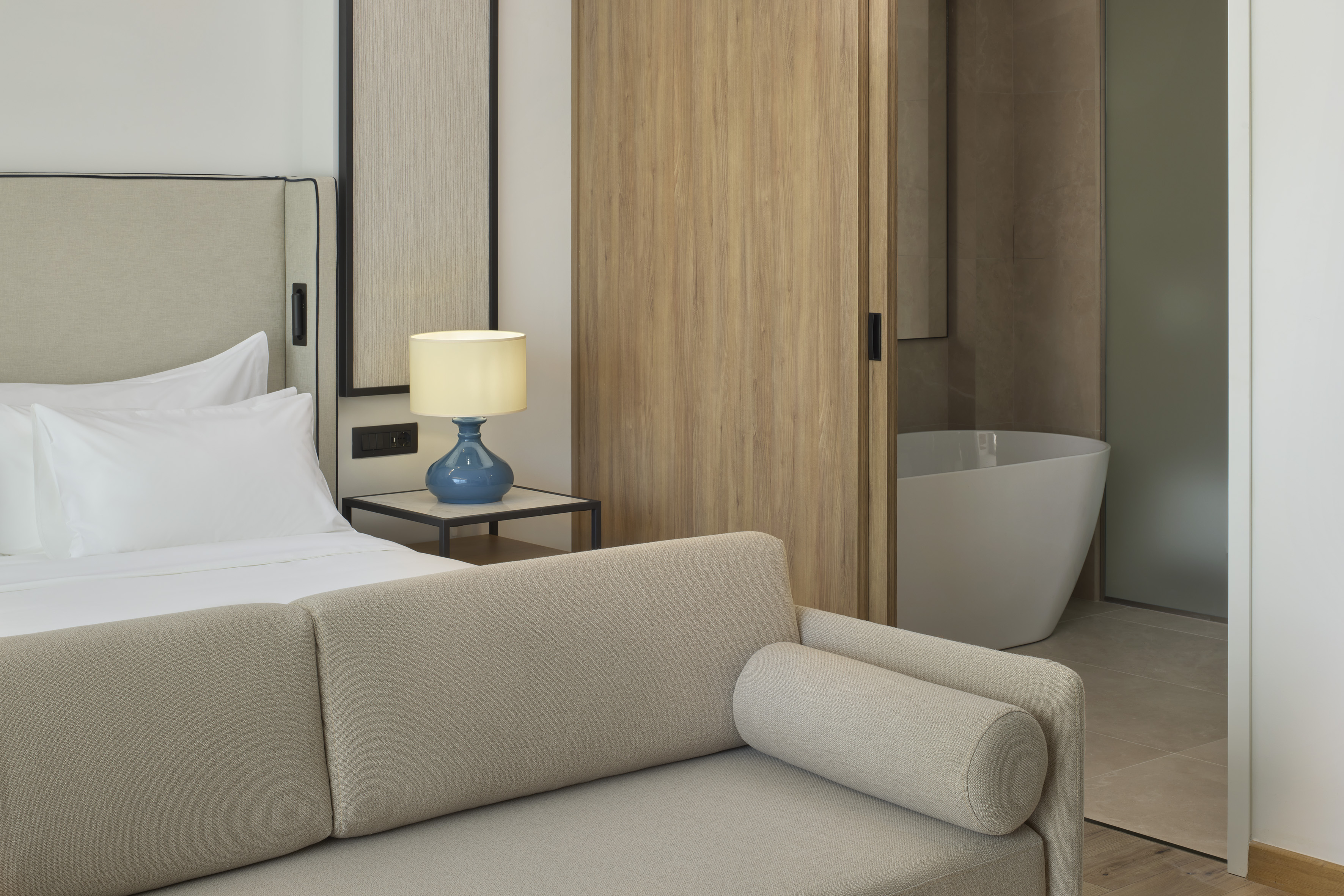 Modern hotel bedroom, sofa, blue lamp. Sliding door to bathroom with tub.