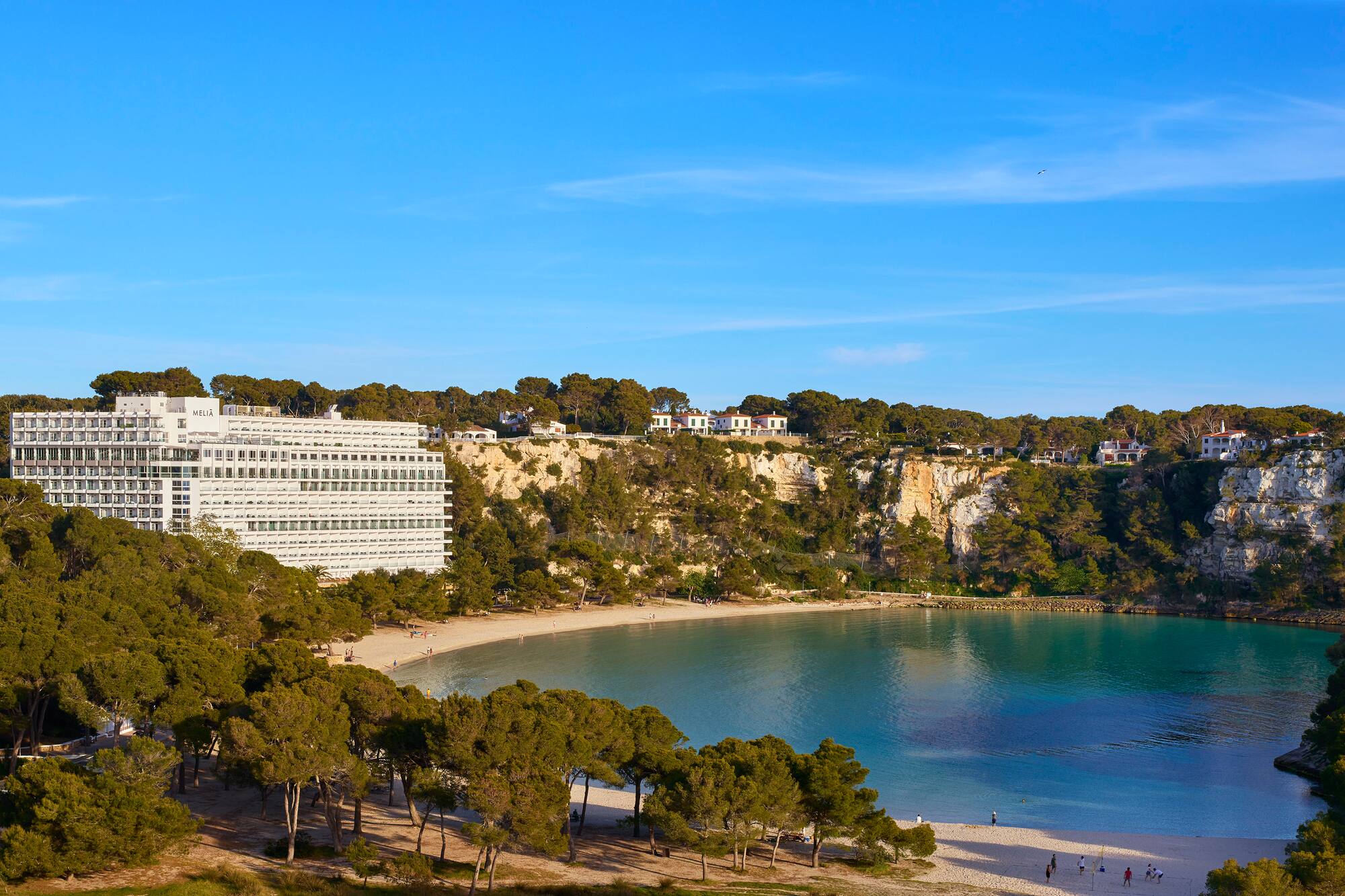 Mallorca beach cove with hotel, turquoise water, pine forests, and rocky cliffs.