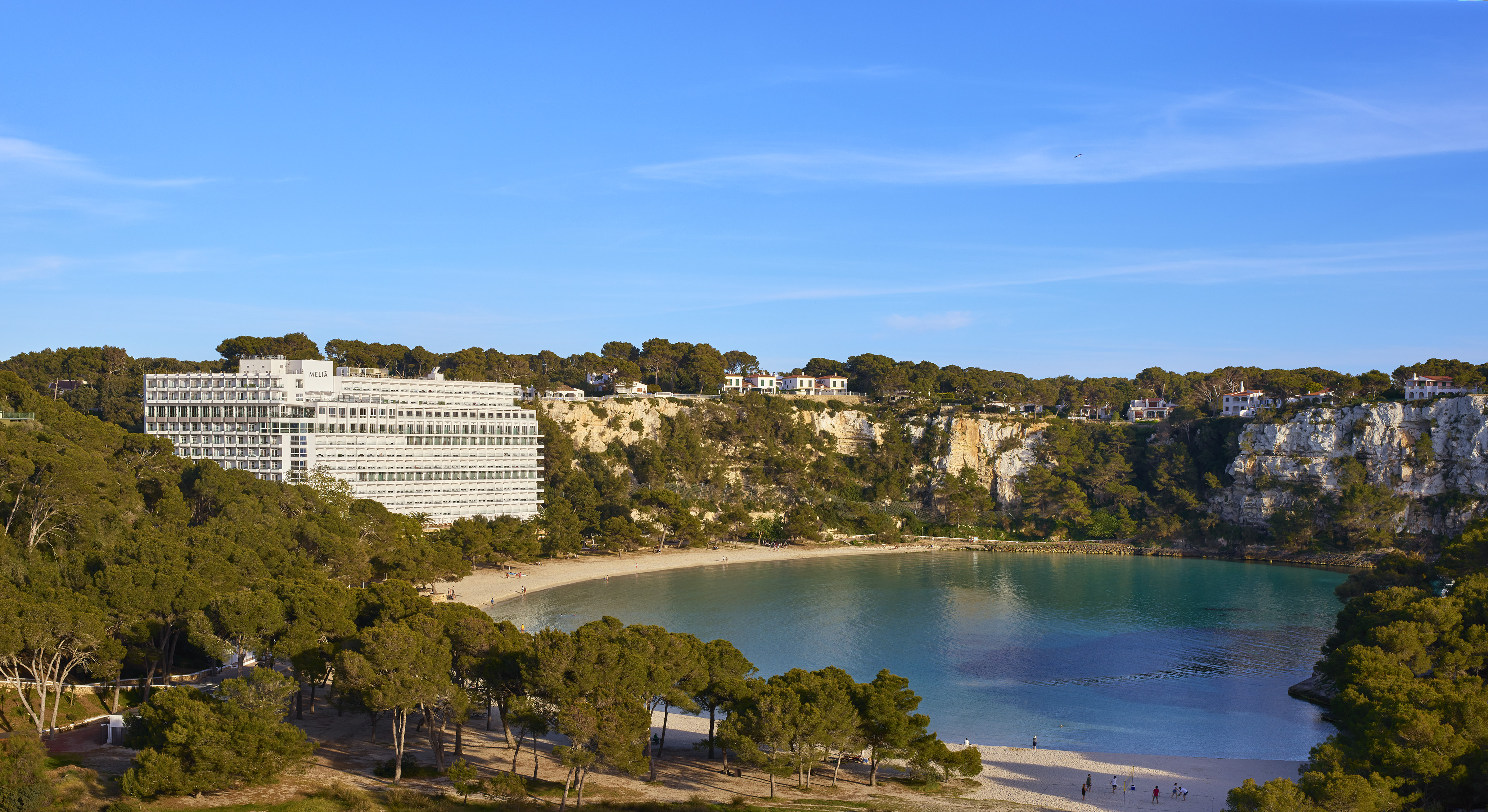 Mallorca beach cove with hotel, turquoise water, pine forests, and rocky cliffs.