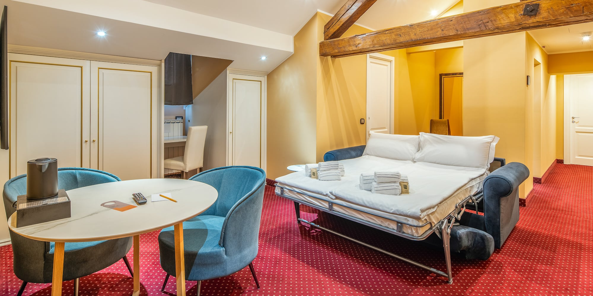 Hotel suite with sofa bed, red patterned carpet, dining area, and wood beams.