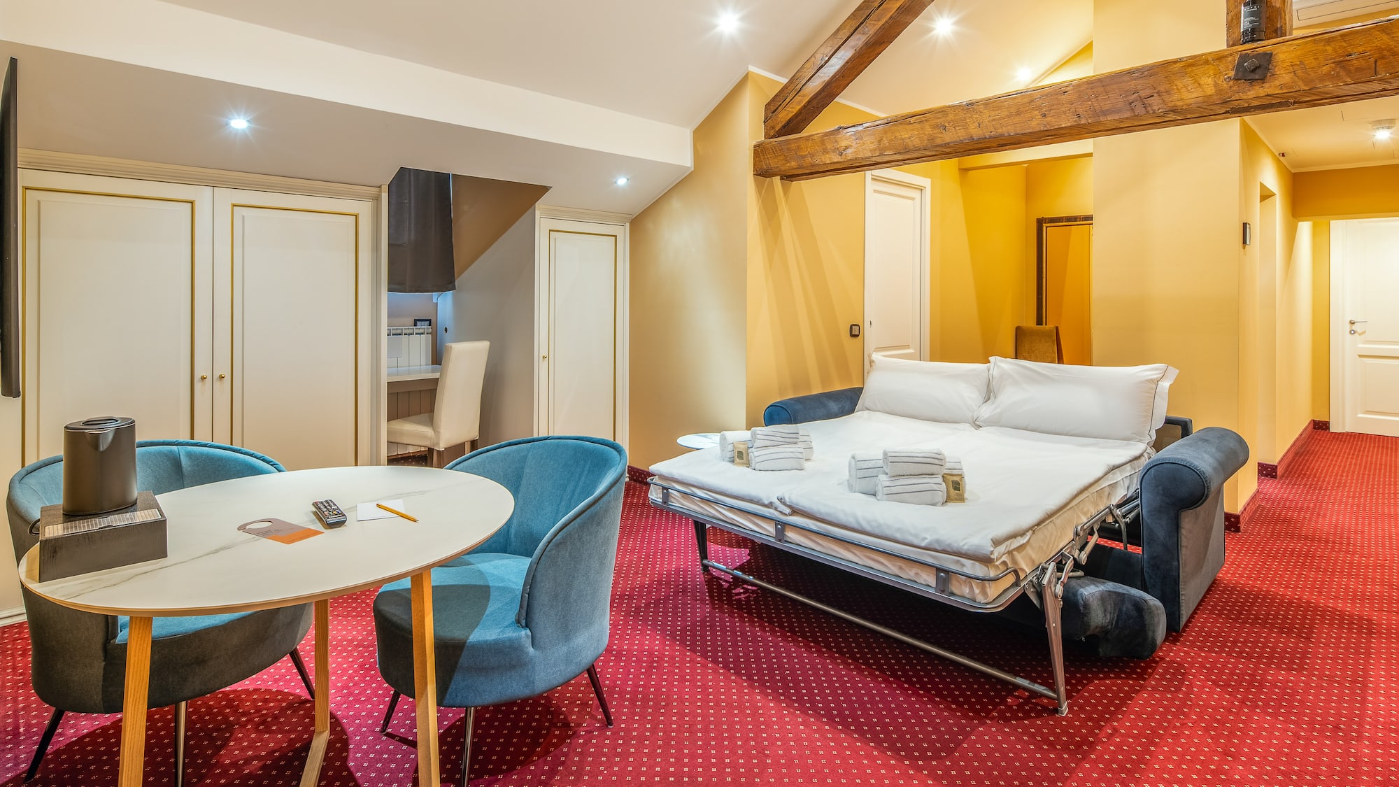 Hotel suite with sofa bed, red patterned carpet, dining area, and wood beams.