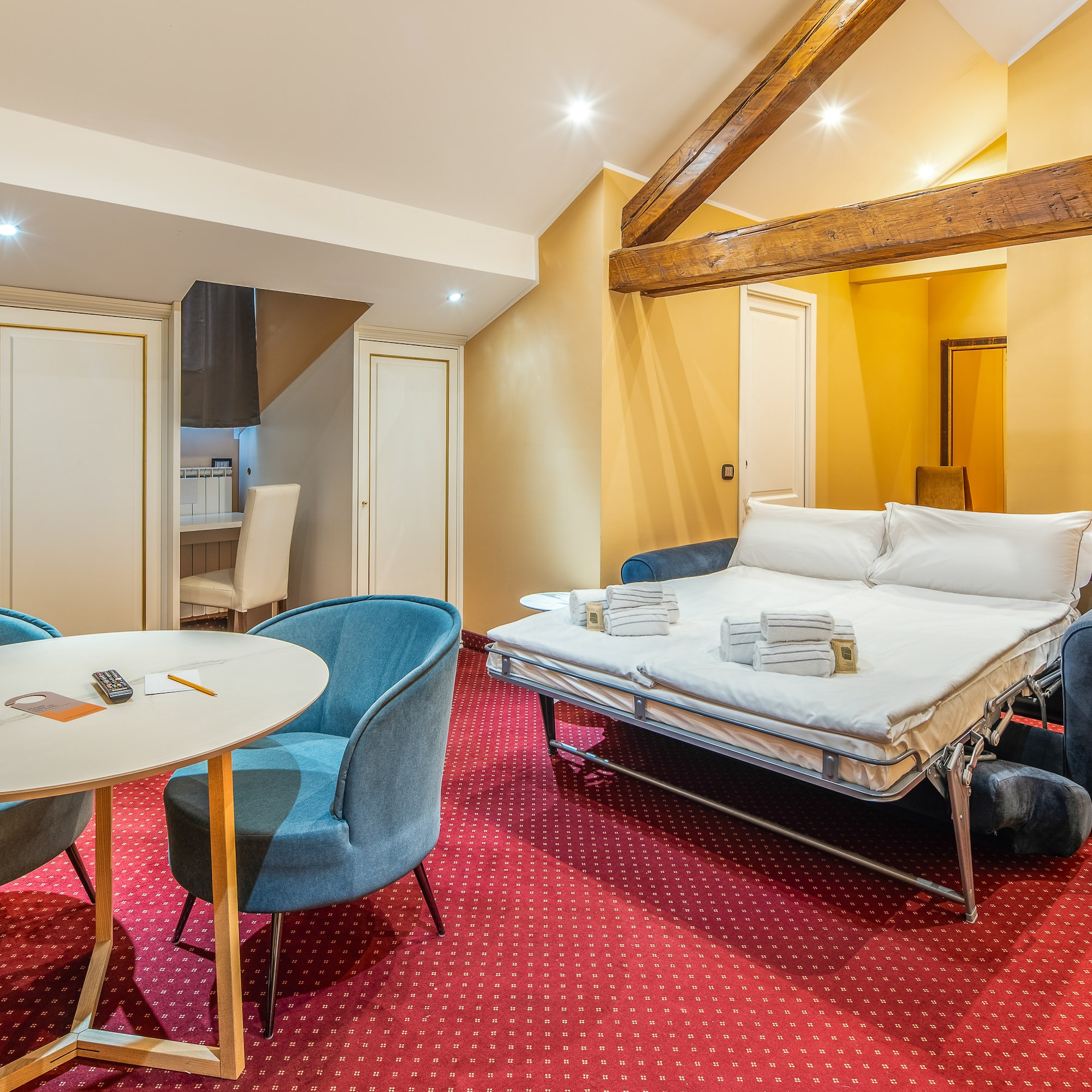 Hotel suite with sofa bed, red patterned carpet, dining area, and wood beams.
