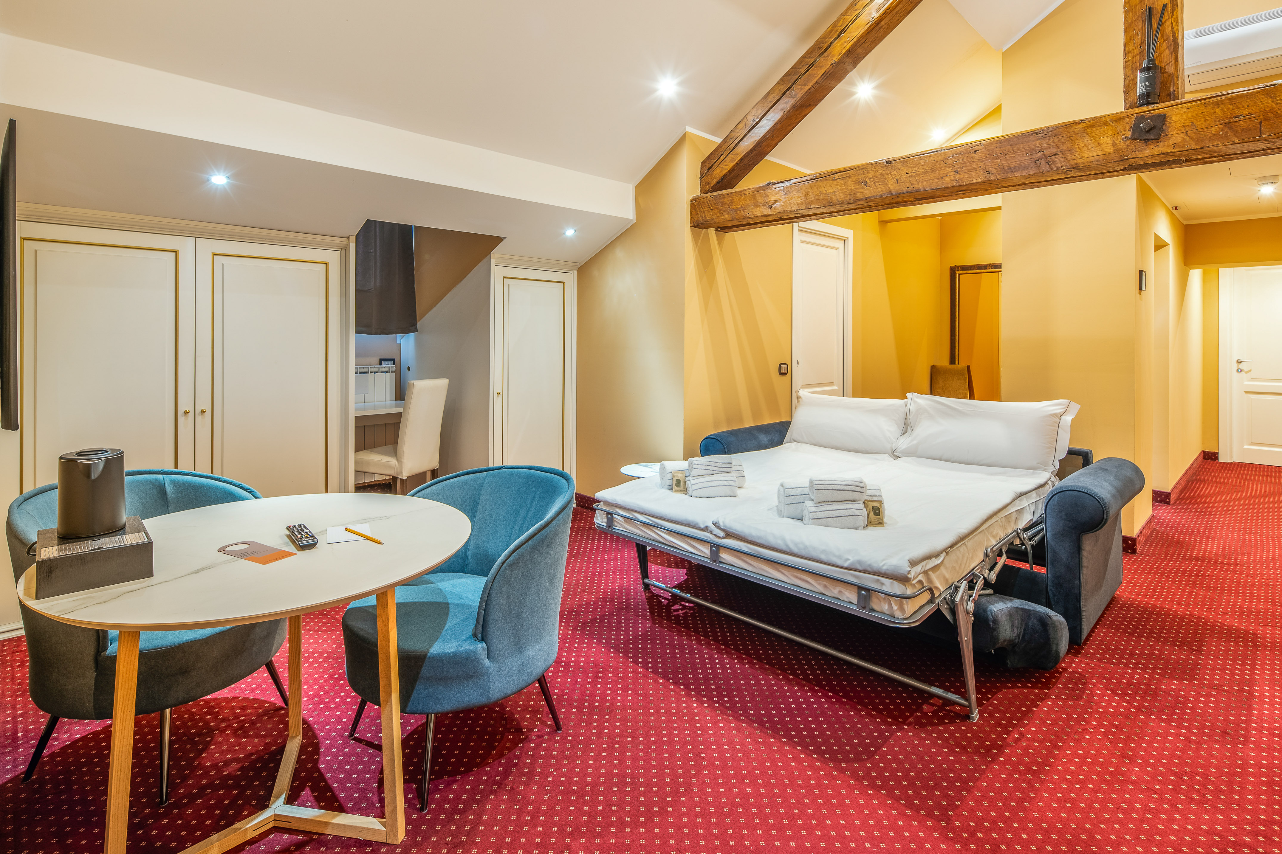 Hotel suite with sofa bed, red patterned carpet, dining area, and wood beams.