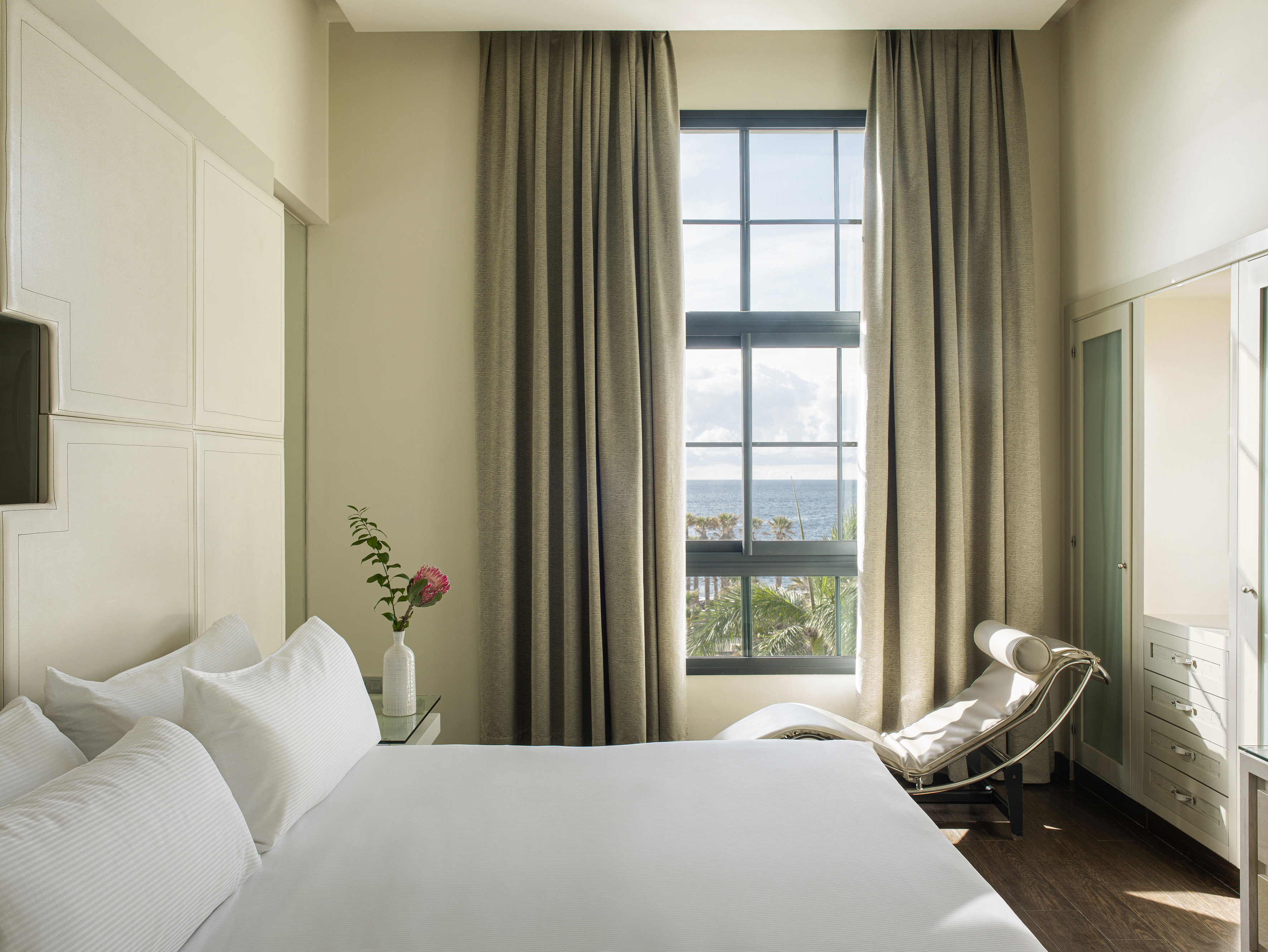 Bright hotel bedroom with ocean view window, white bed, and modern chaise.
