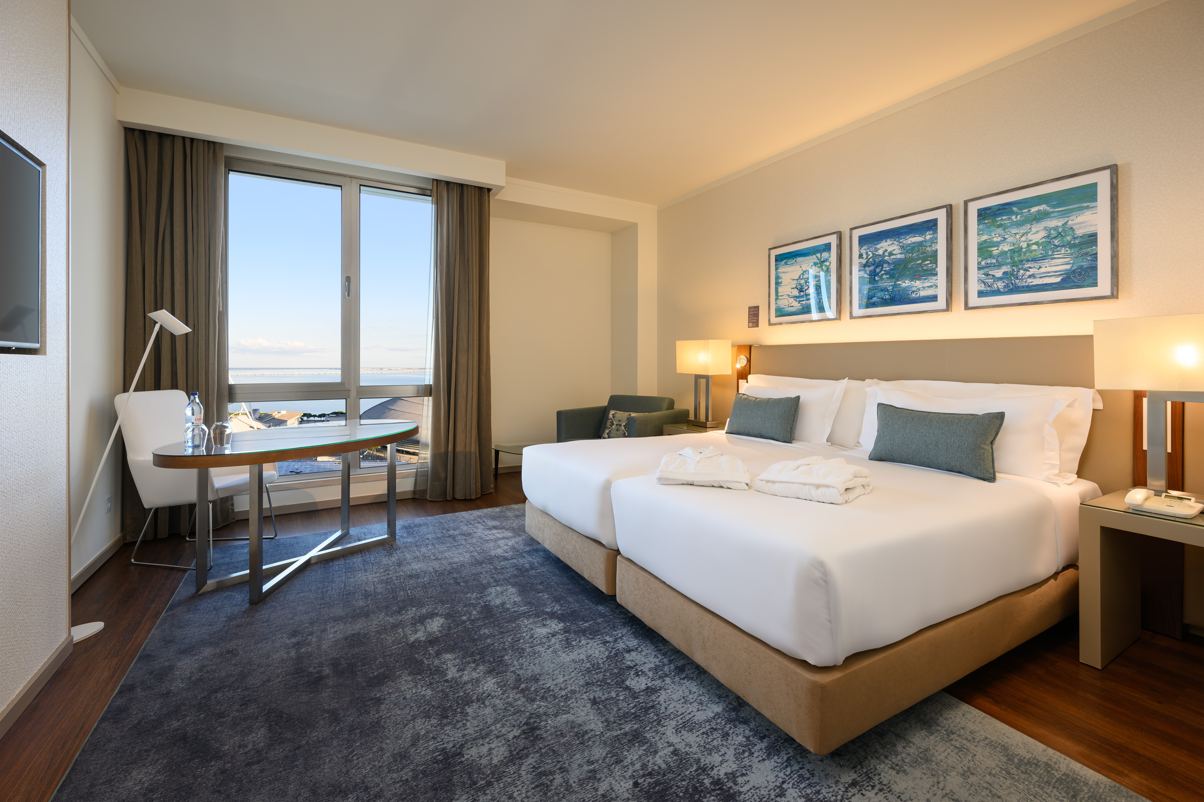 Modern hotel room with twin beds, waterfront view, desk, and abstract wall art.