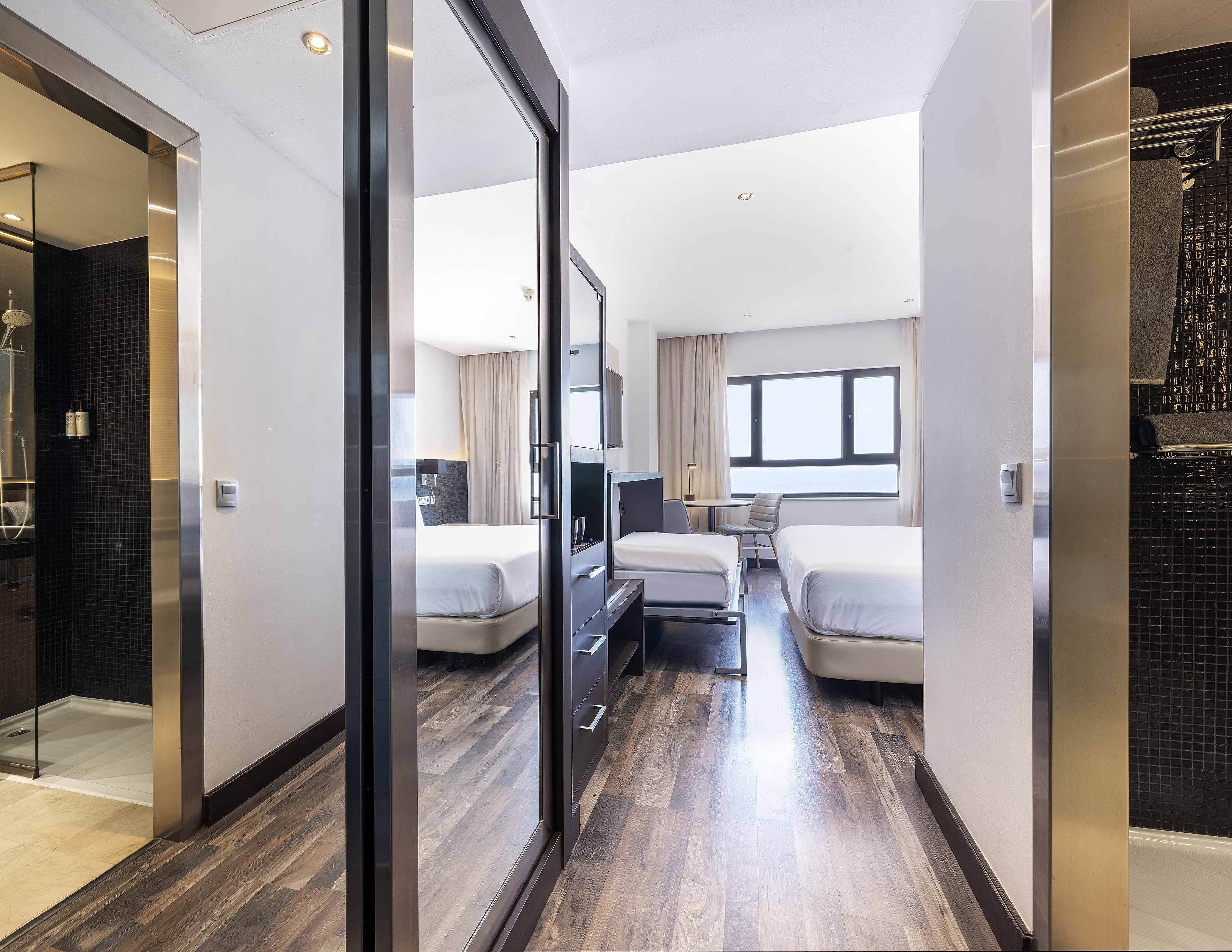 Modern hotel room with twin beds, wooden floor, window, and glass shower.