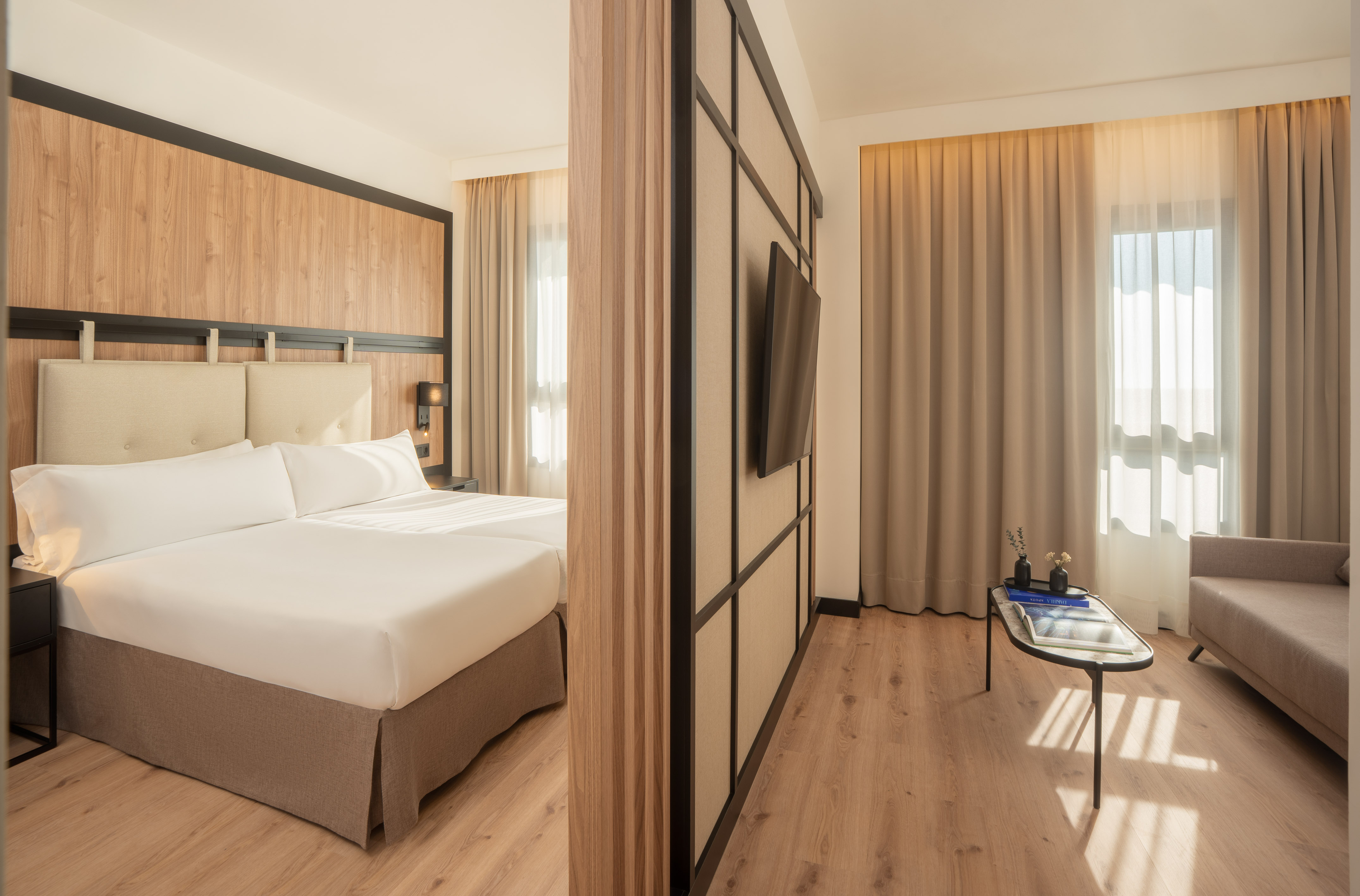 Modern hotel suite: twin beds, wood partition, TV, living area with sofa.