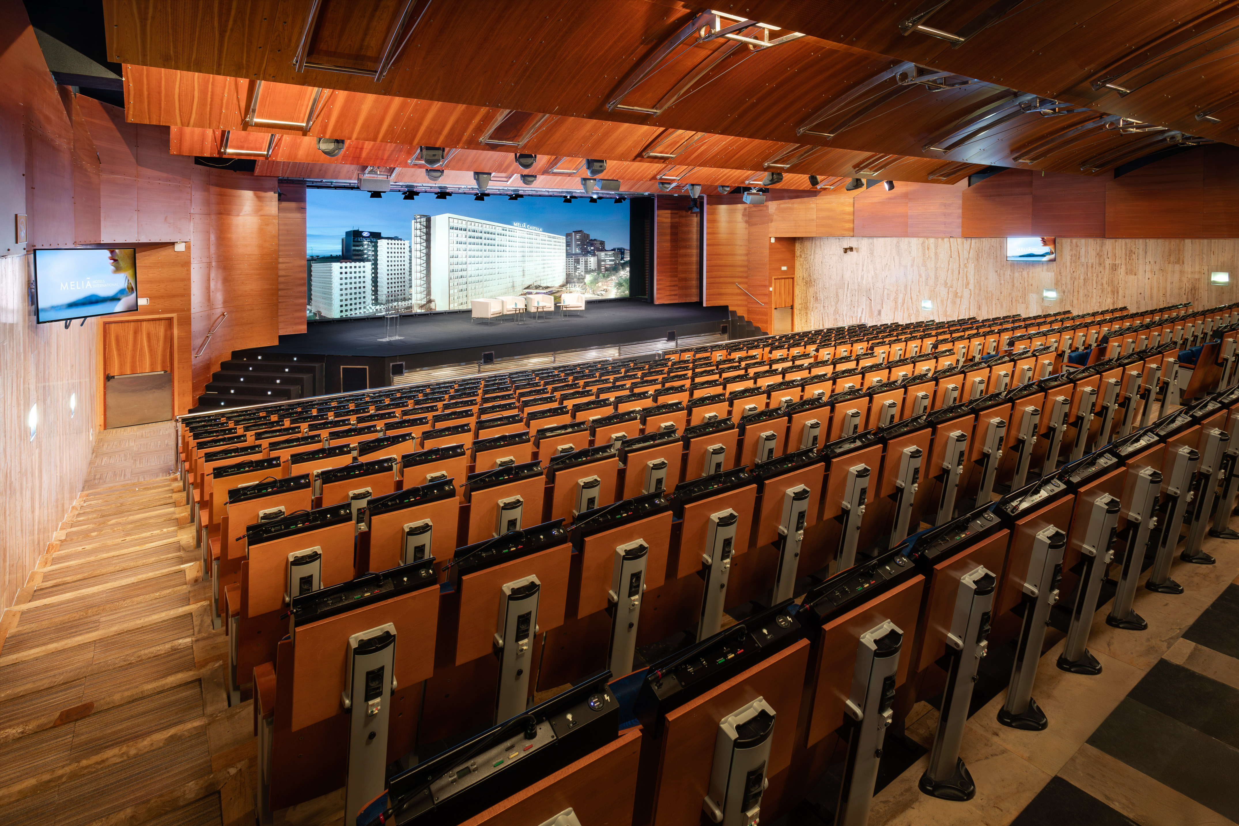 Modern auditorium with tiered seating, stage, and large screen display.