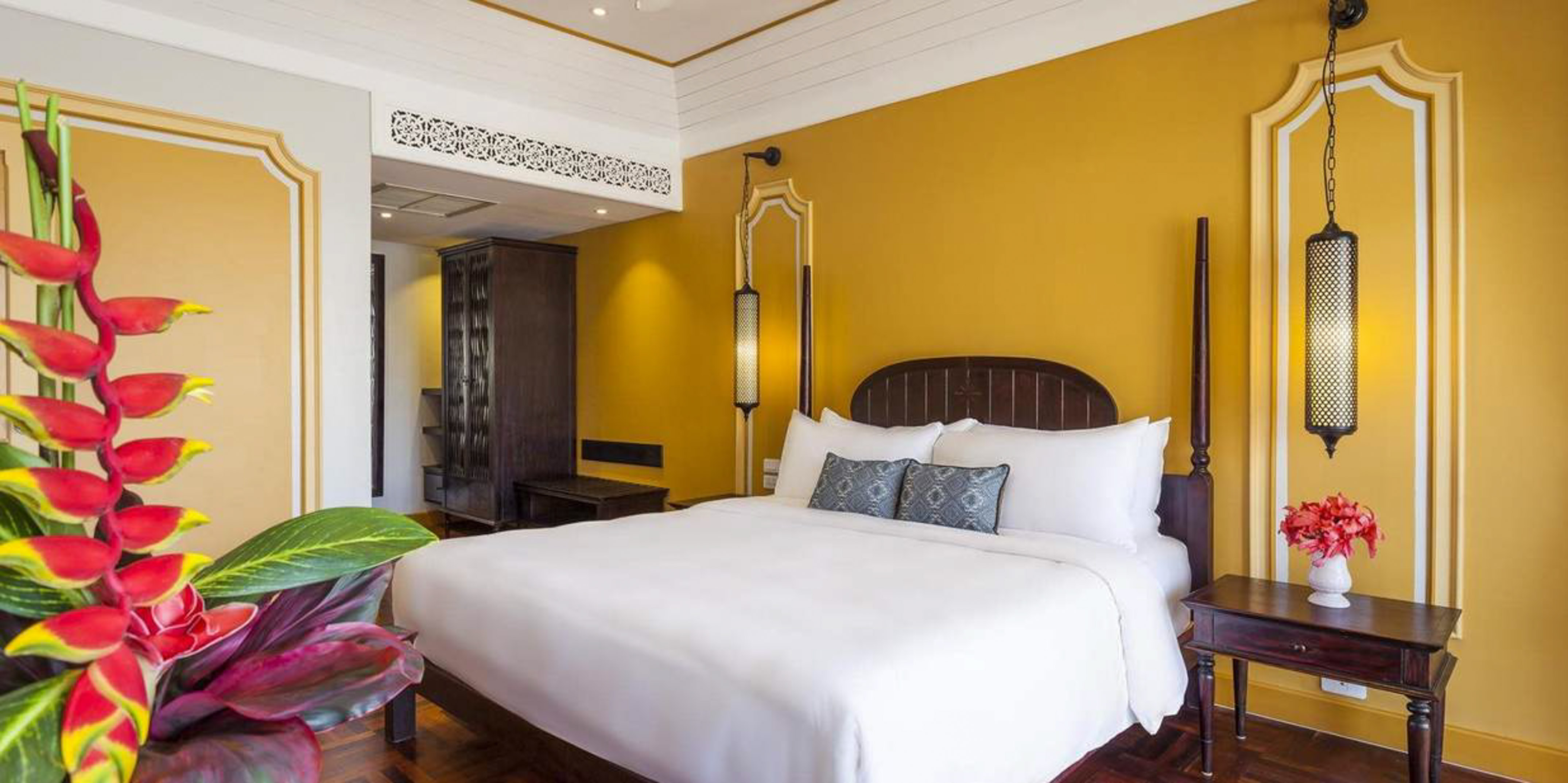 Hotel bedroom with mustard yellow walls, king bed, and tropical flowers.