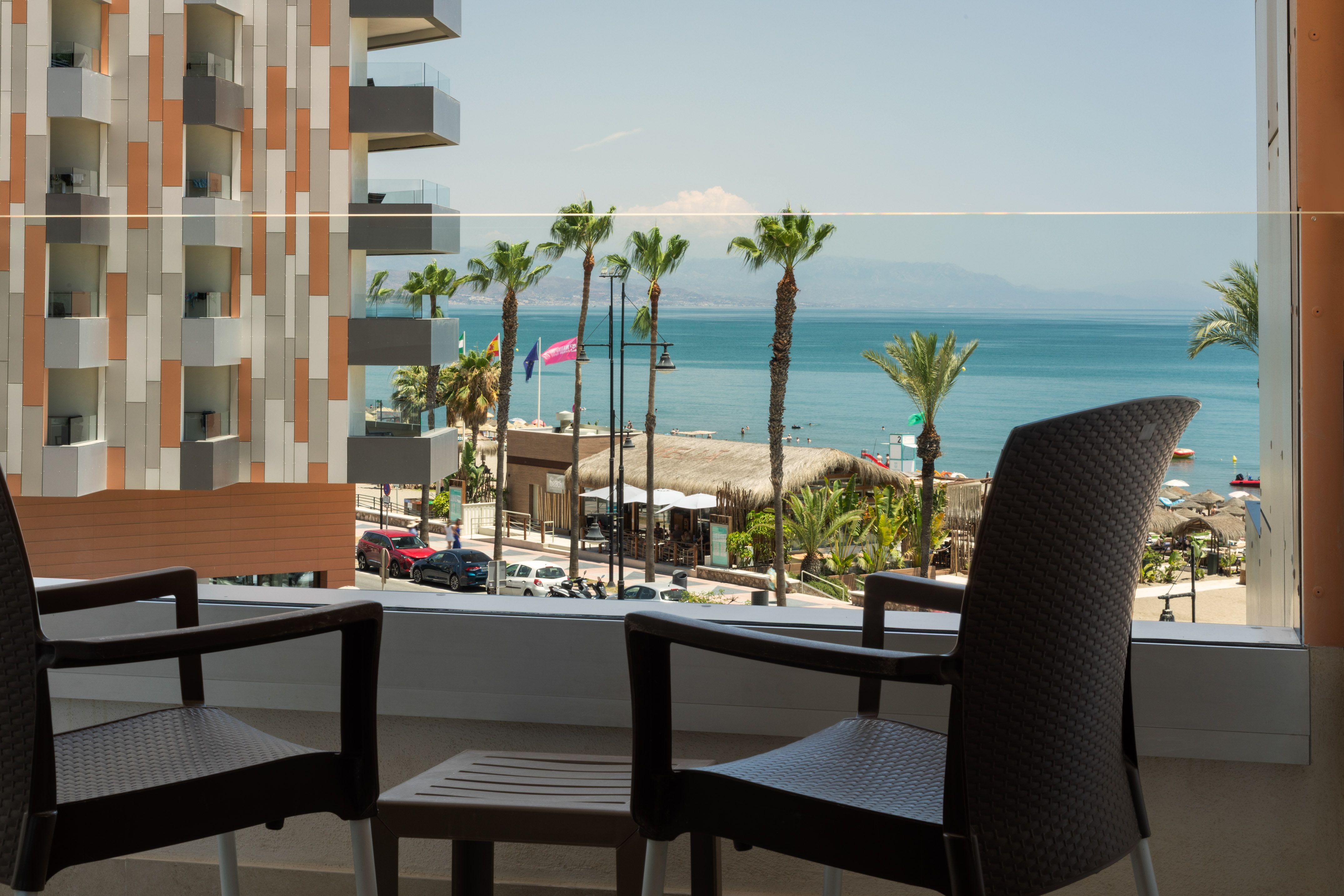 Modern hotel balcony overlooking a sunny beach, palm trees, and blue ocean.