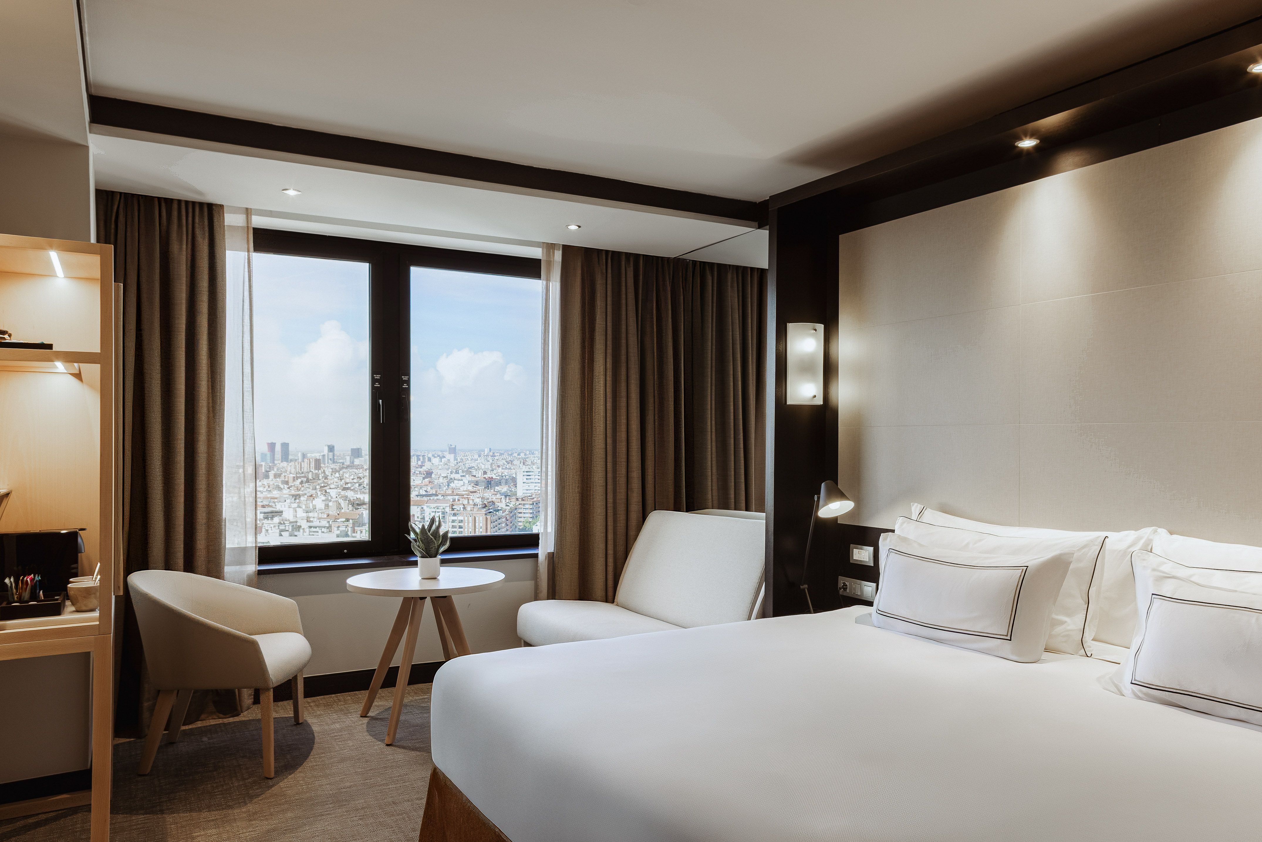 Modern hotel room with city view, white bed, and armchair by the window.