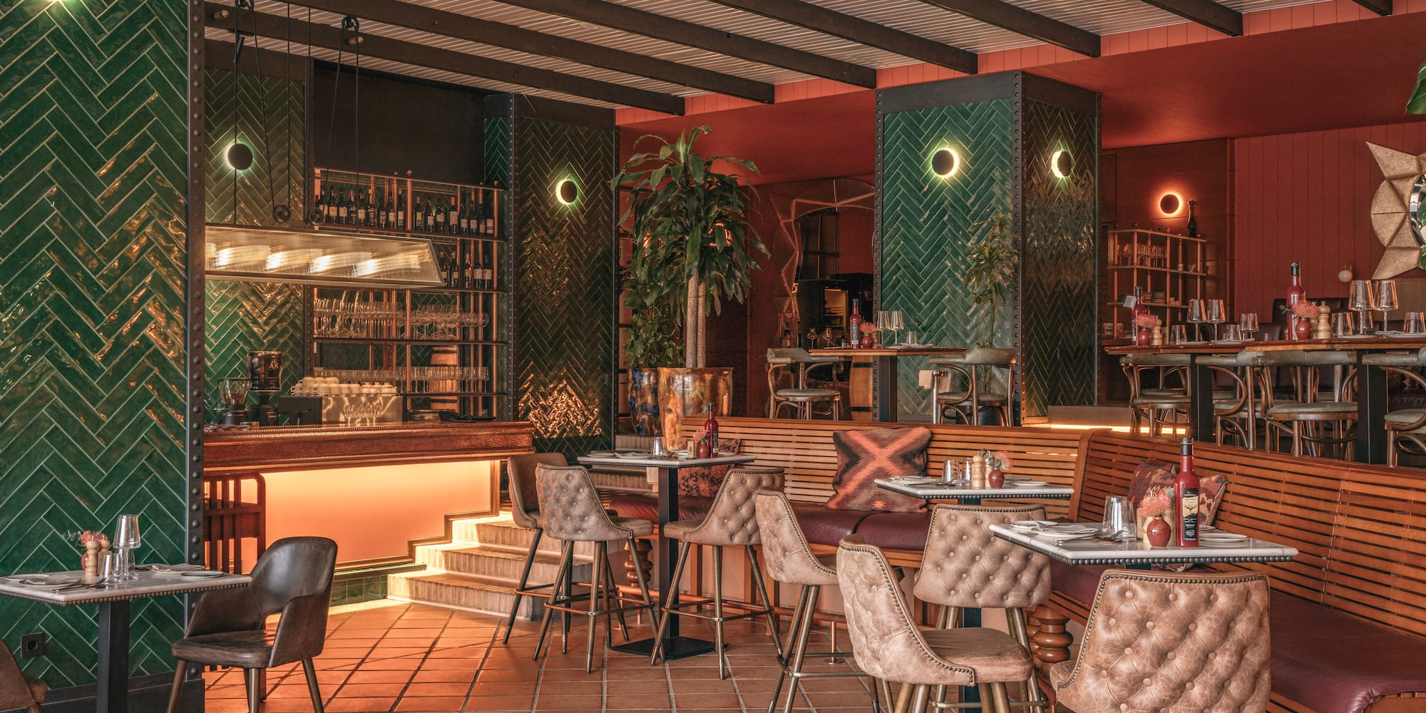 Modern restaurant interior with green herringbone tiles, warm lighting, bar, and terracotta floor.