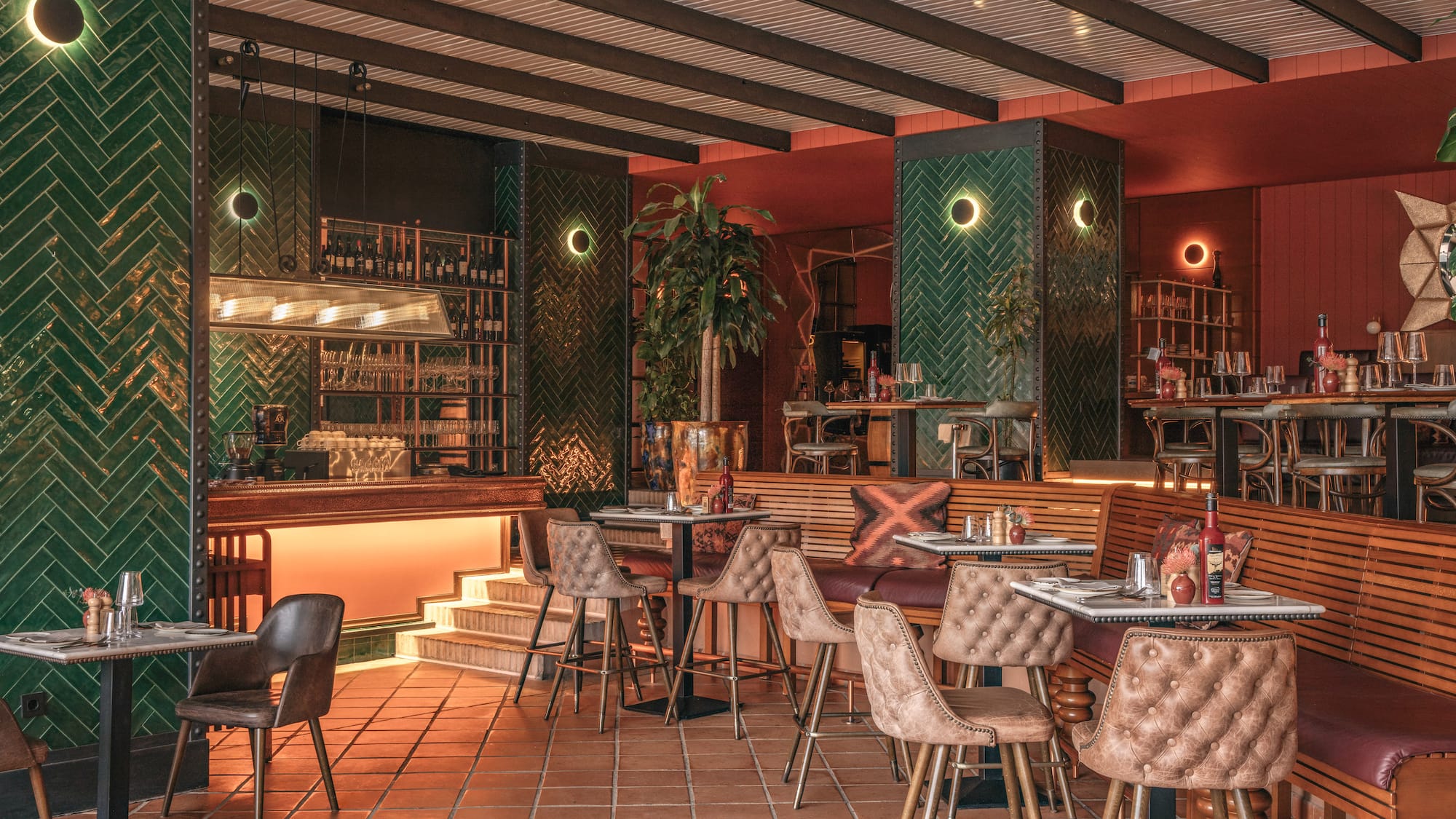 Modern restaurant interior with green herringbone tiles, warm lighting, bar, and terracotta floor.