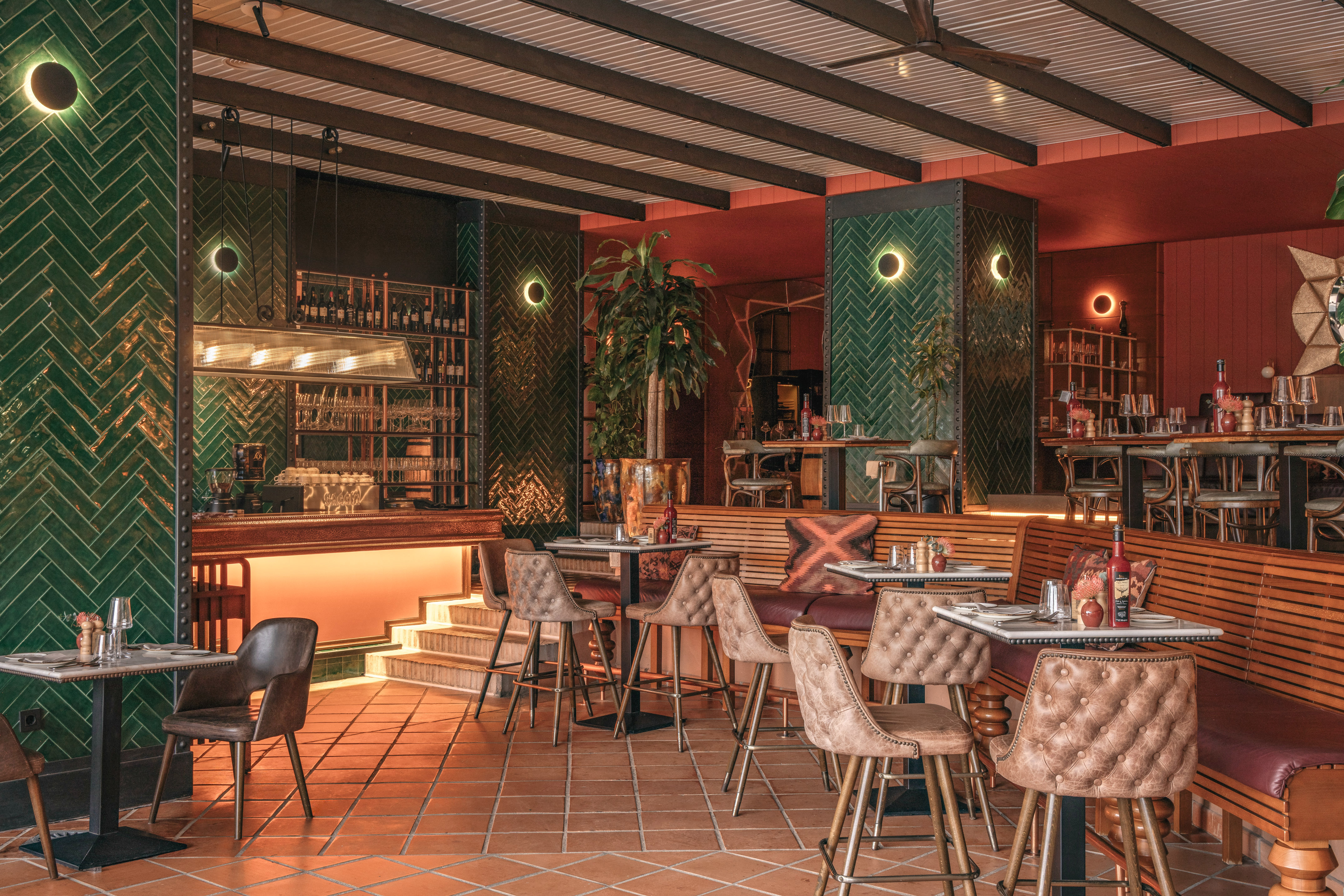 Modern restaurant interior with green herringbone tiles, warm lighting, bar, and terracotta floor.