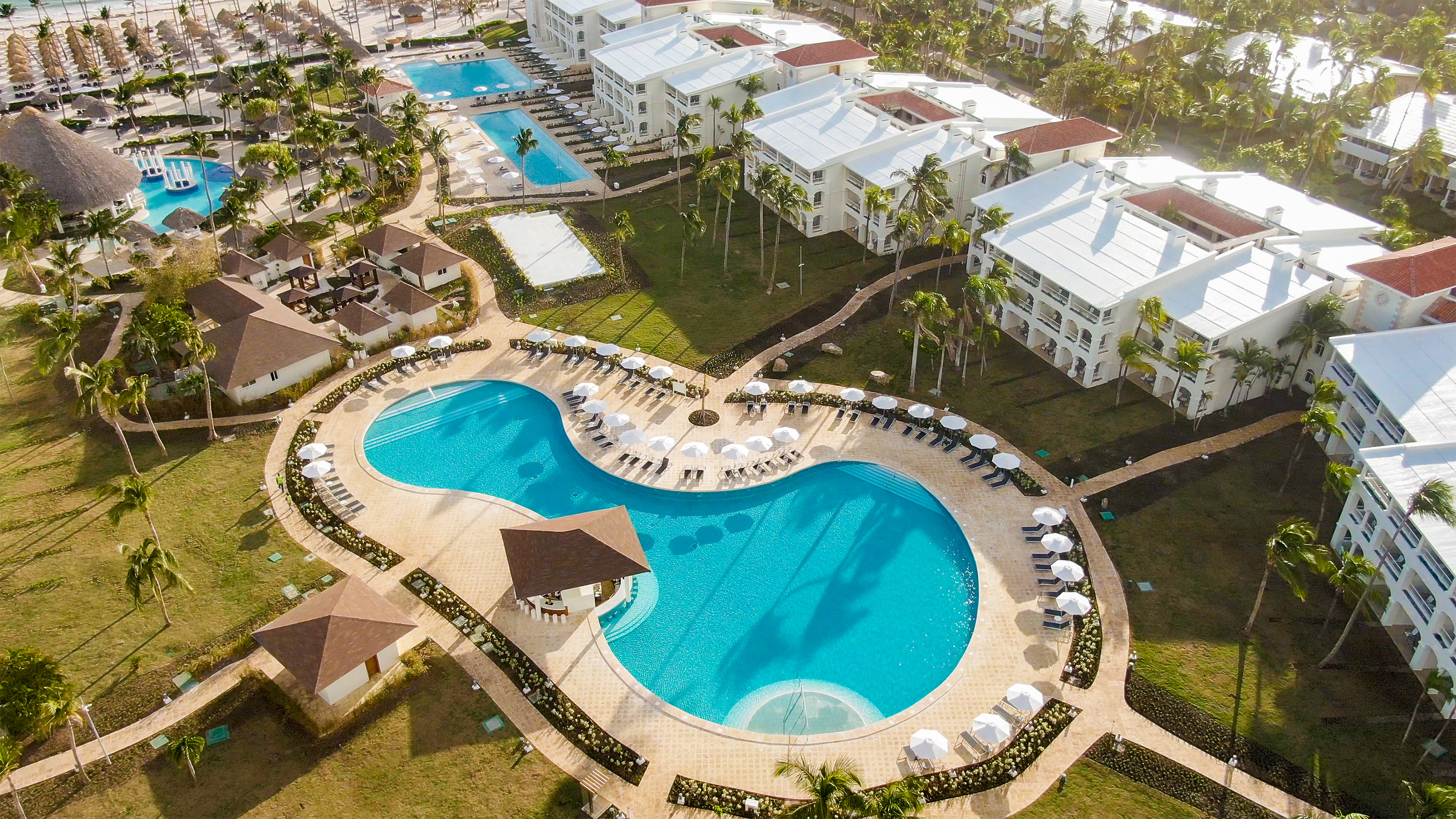 Aerial view: tropical resort pool, swim-up bar, white hotels, lush palm trees.