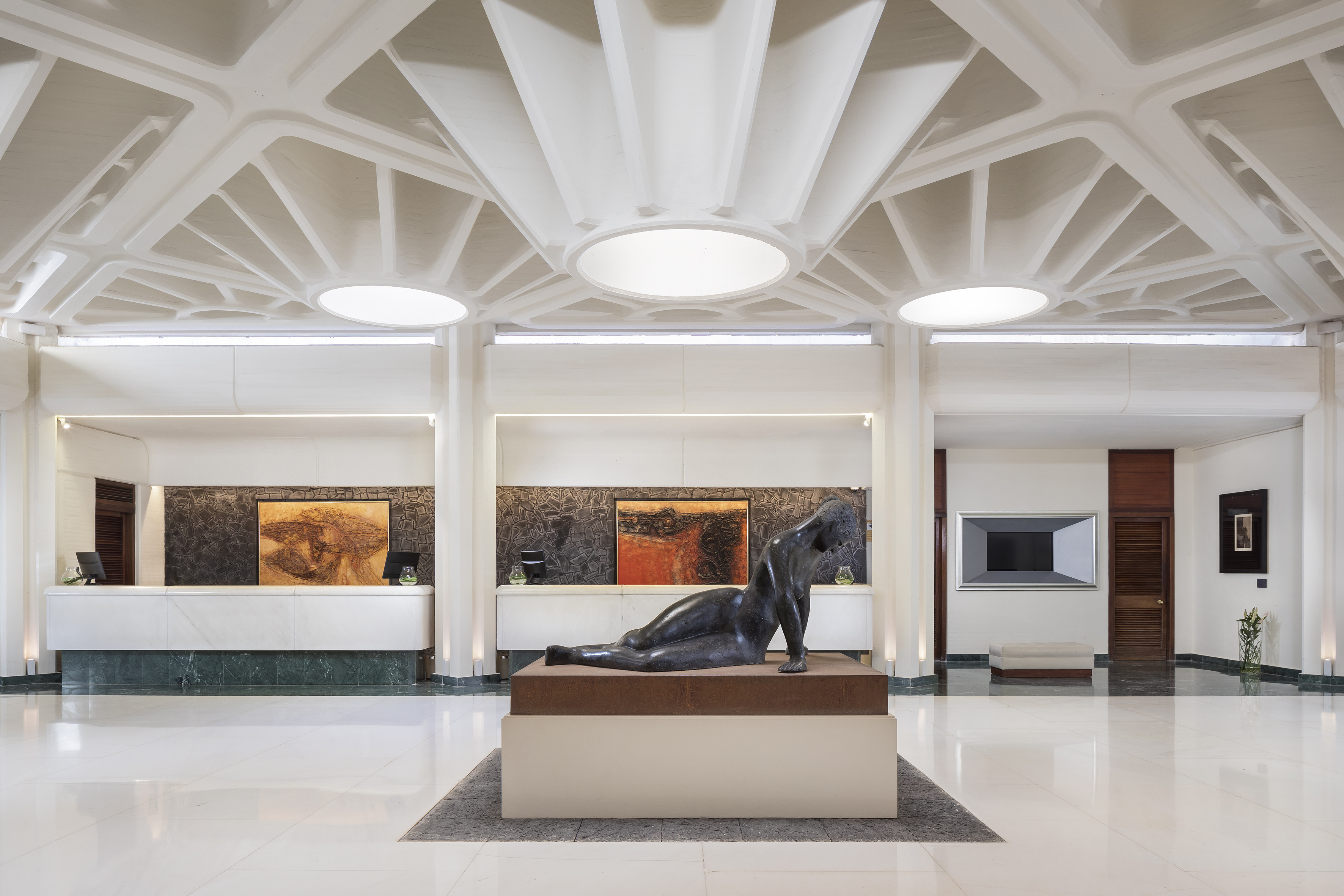 Modern hotel lobby with geometric ceiling, reception area, and central reclining sculpture.