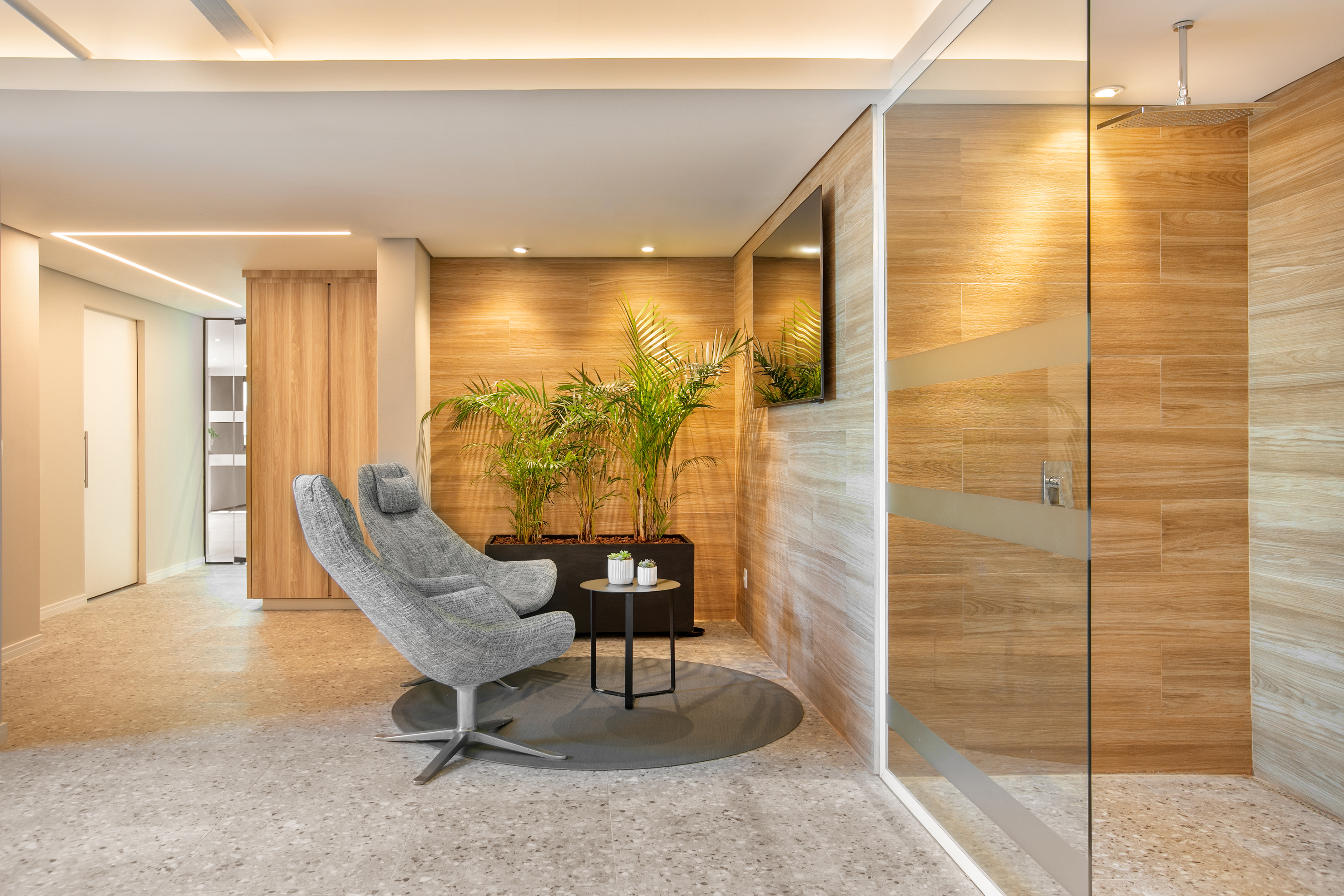 Modern lounge area with gray recliner, green plants, wood wall, and glass shower.