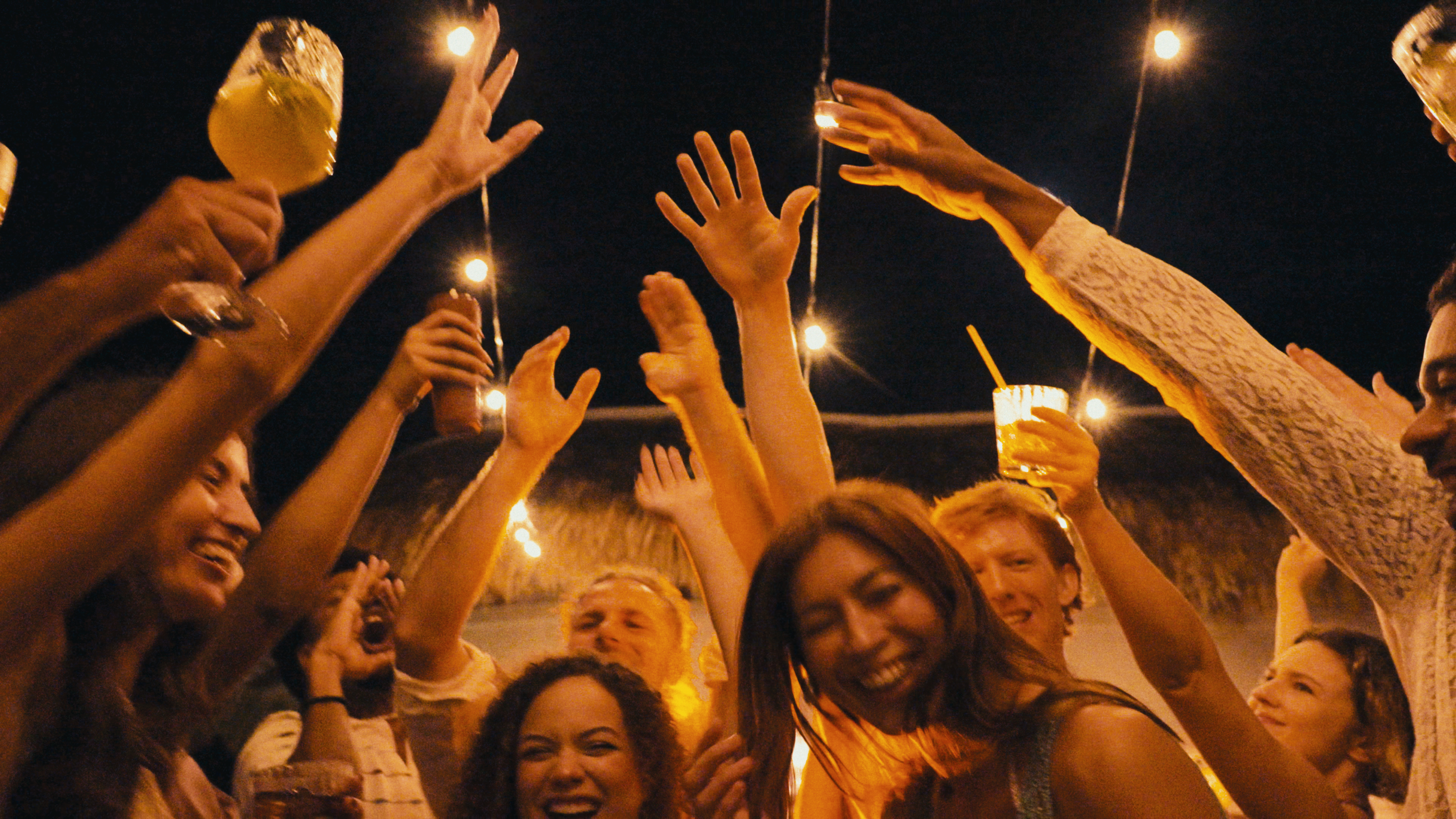 Joyful partygoers raising hands and drinks under festive string lights at night.