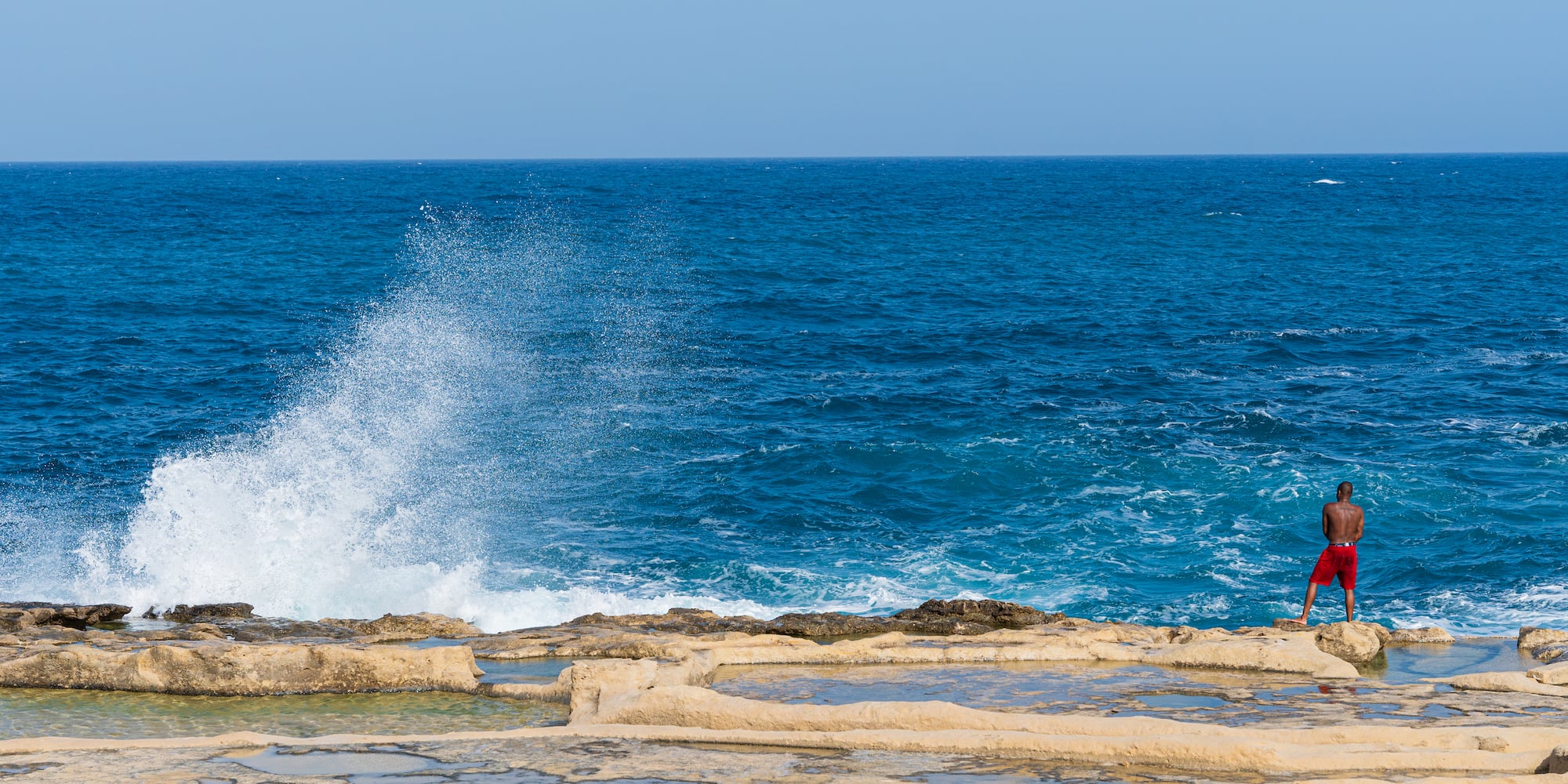 Man on rocky coast by blue ocean with salt pans, watching wave.