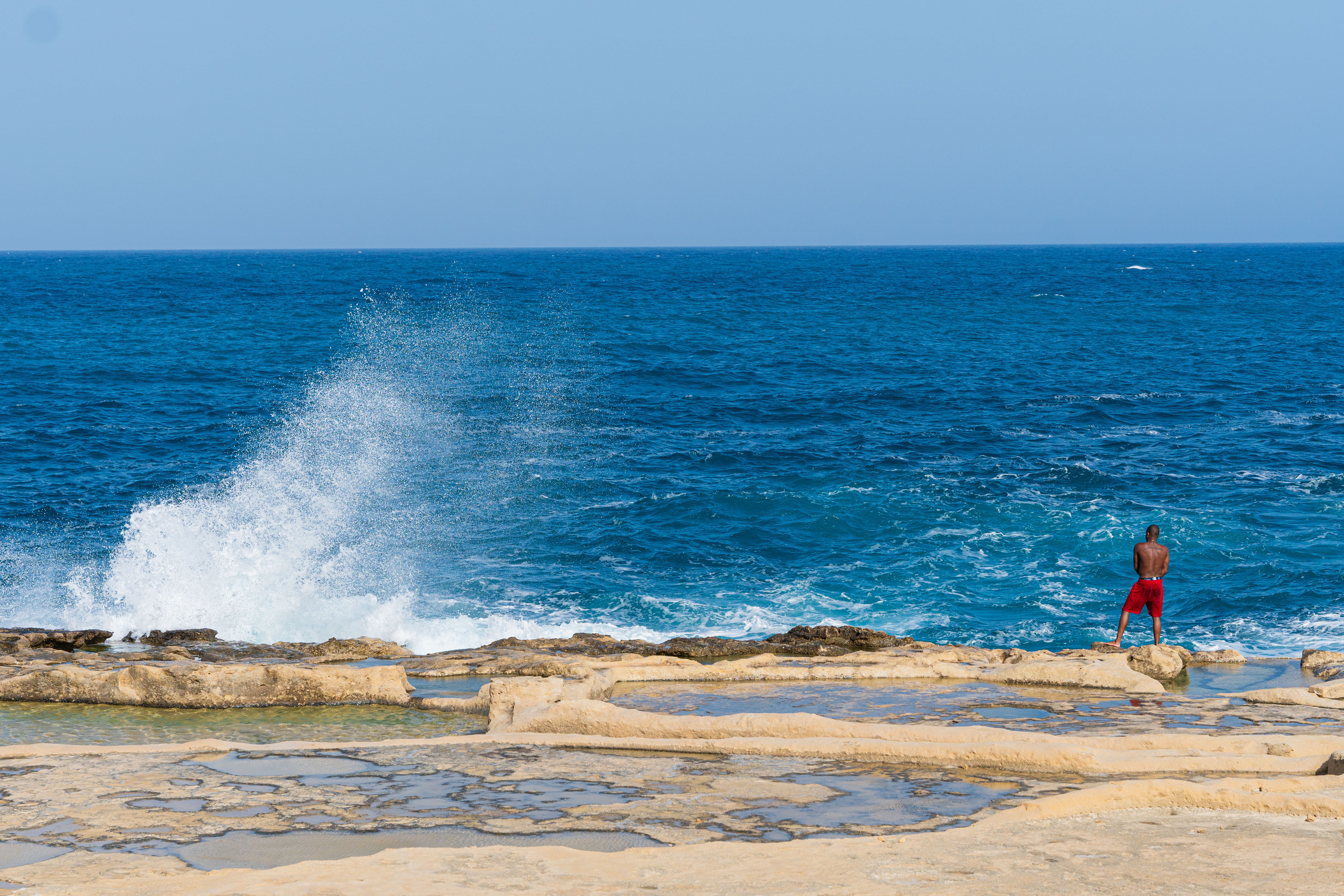 Man on rocky coast by blue ocean with salt pans, watching wave.
