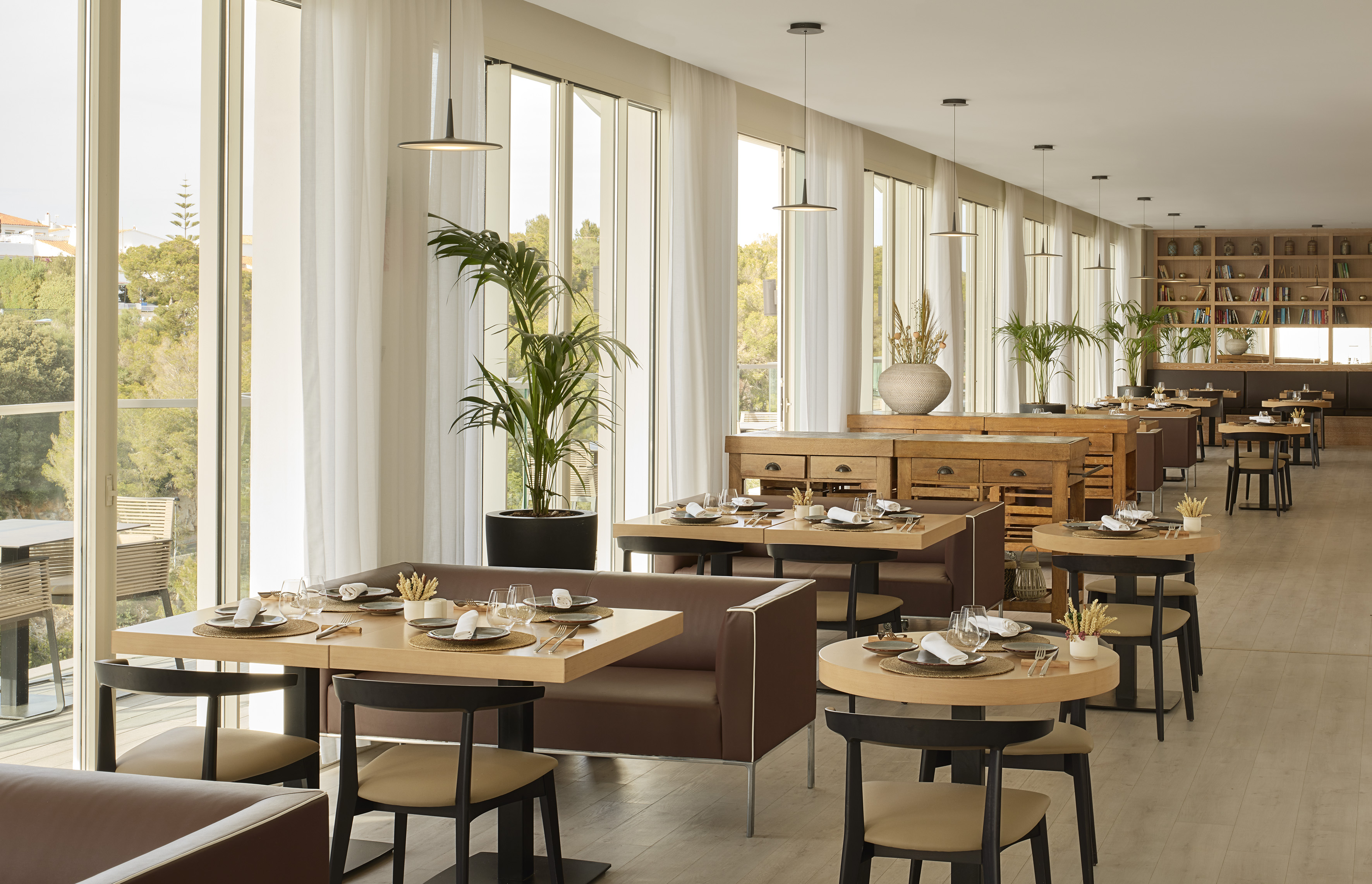 Modern restaurant with wooden tables, brown booths, plants, and bright window views.