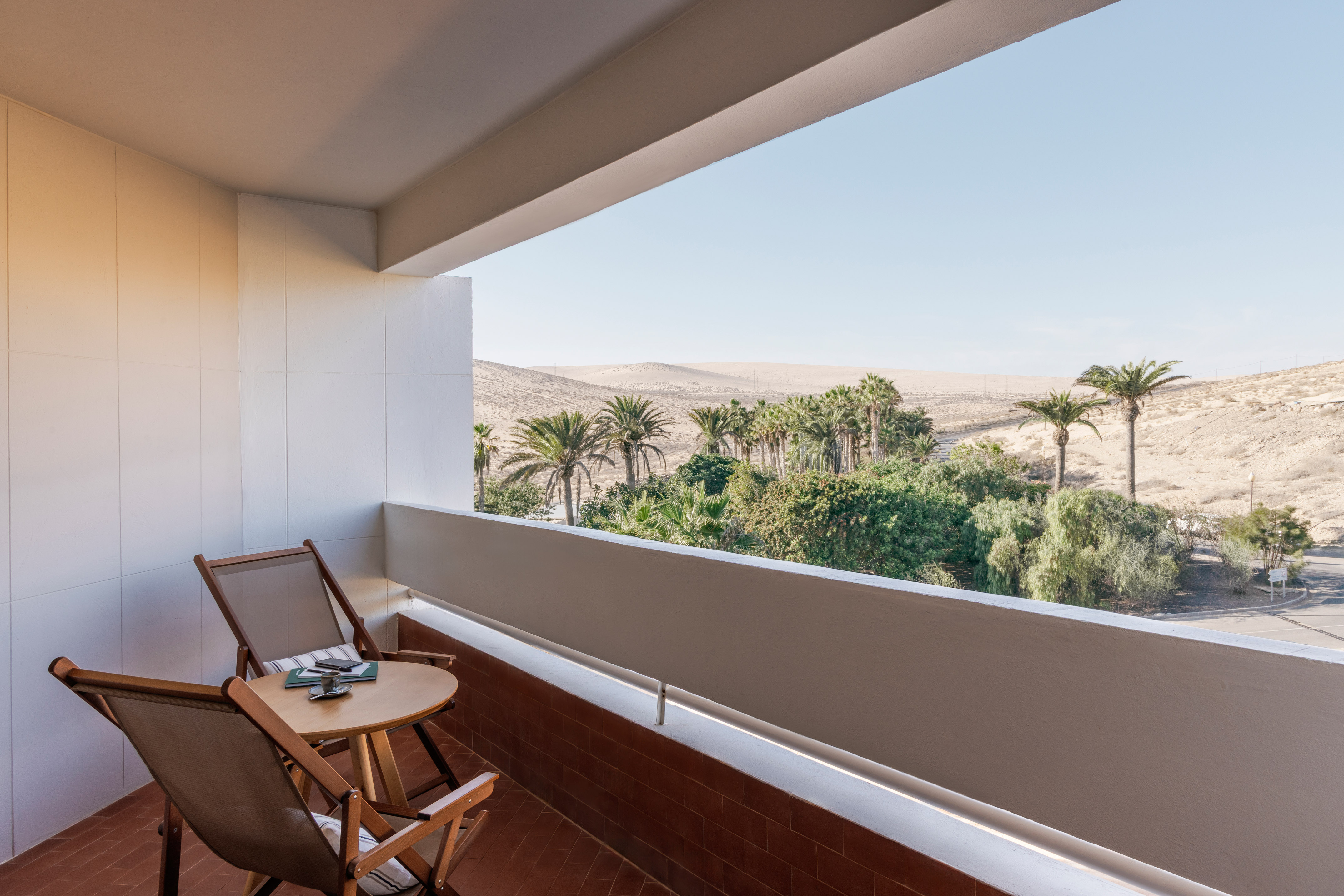 Hotel balcony with chairs, palm trees, and desert landscape view.