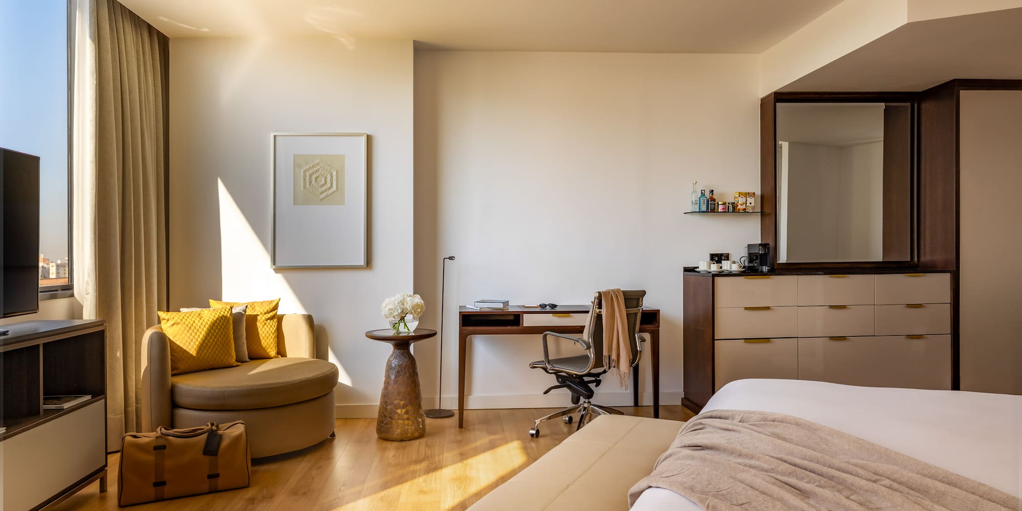 Sunny modern hotel room with bed, work desk, armchair, and city view.