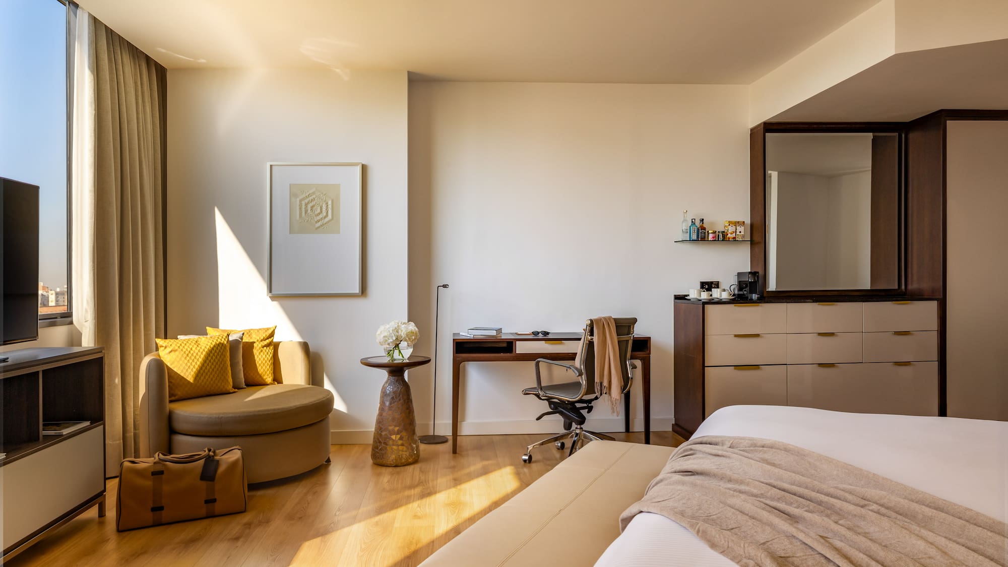 Sunny modern hotel room with bed, work desk, armchair, and city view.