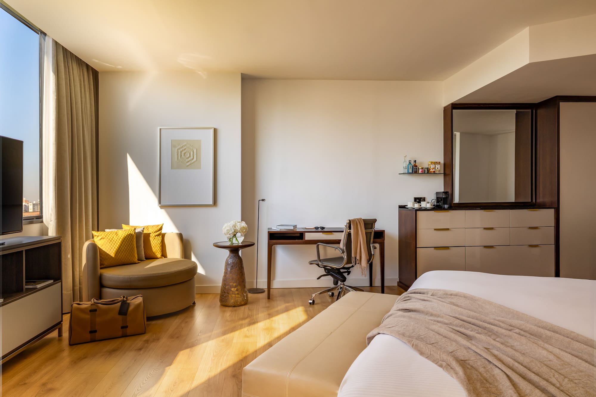 Sunny modern hotel room with bed, work desk, armchair, and city view.