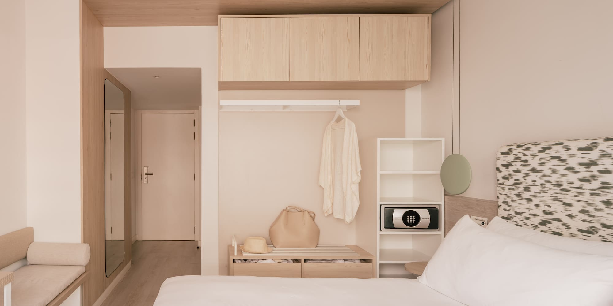 Minimalist hotel bedroom with wood storage, white bed, and in-room safe.