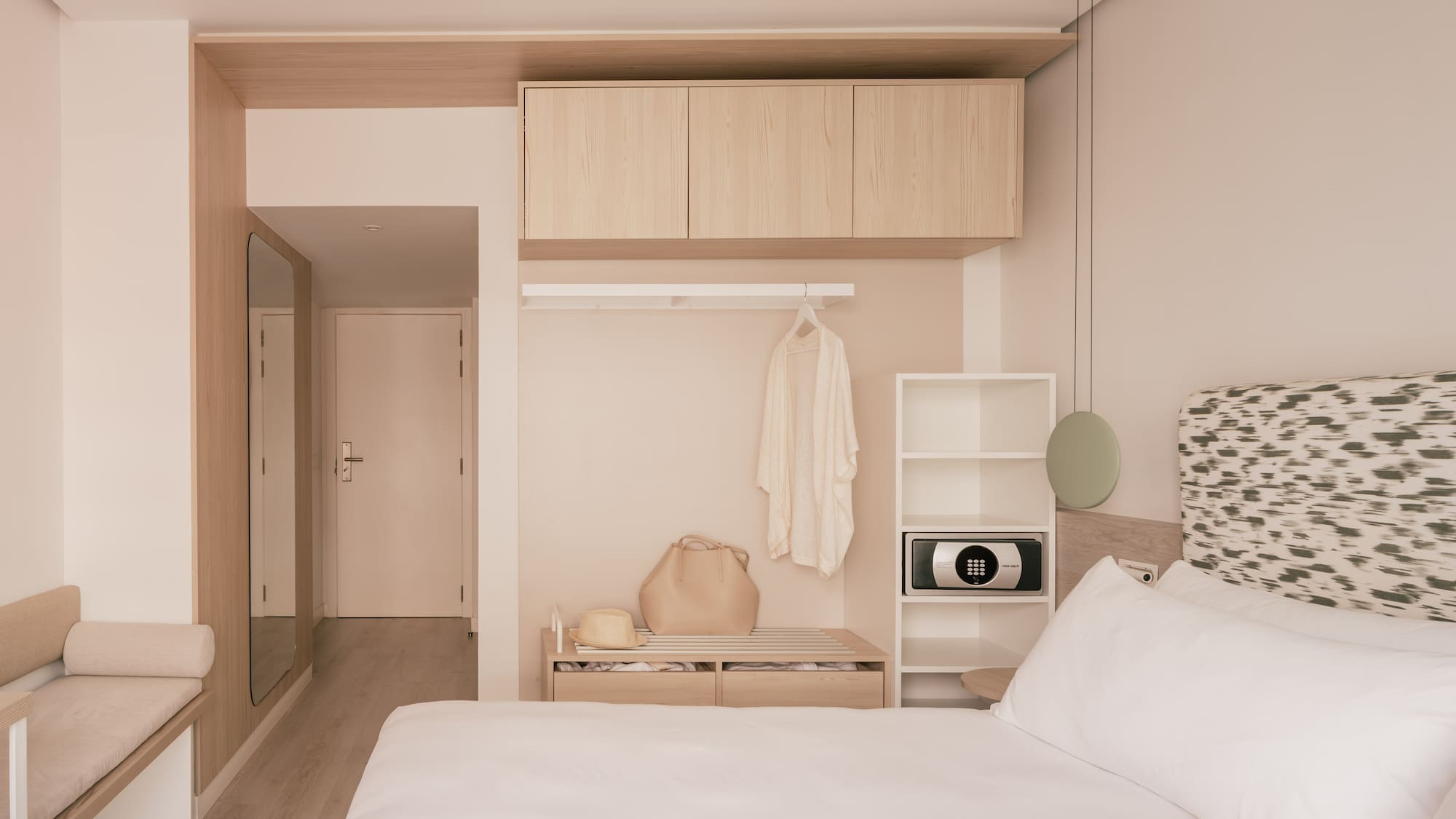 Minimalist hotel bedroom with wood storage, white bed, and in-room safe.