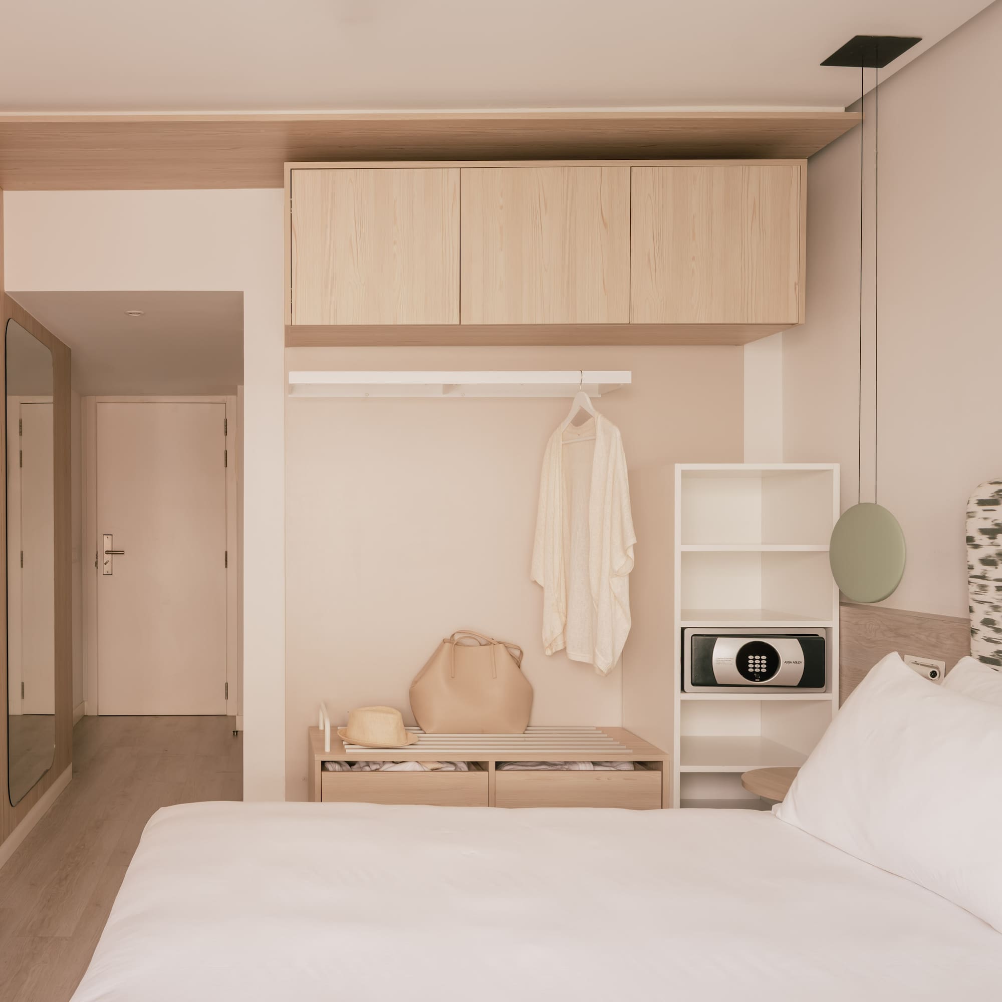 Minimalist hotel bedroom with wood storage, white bed, and in-room safe.