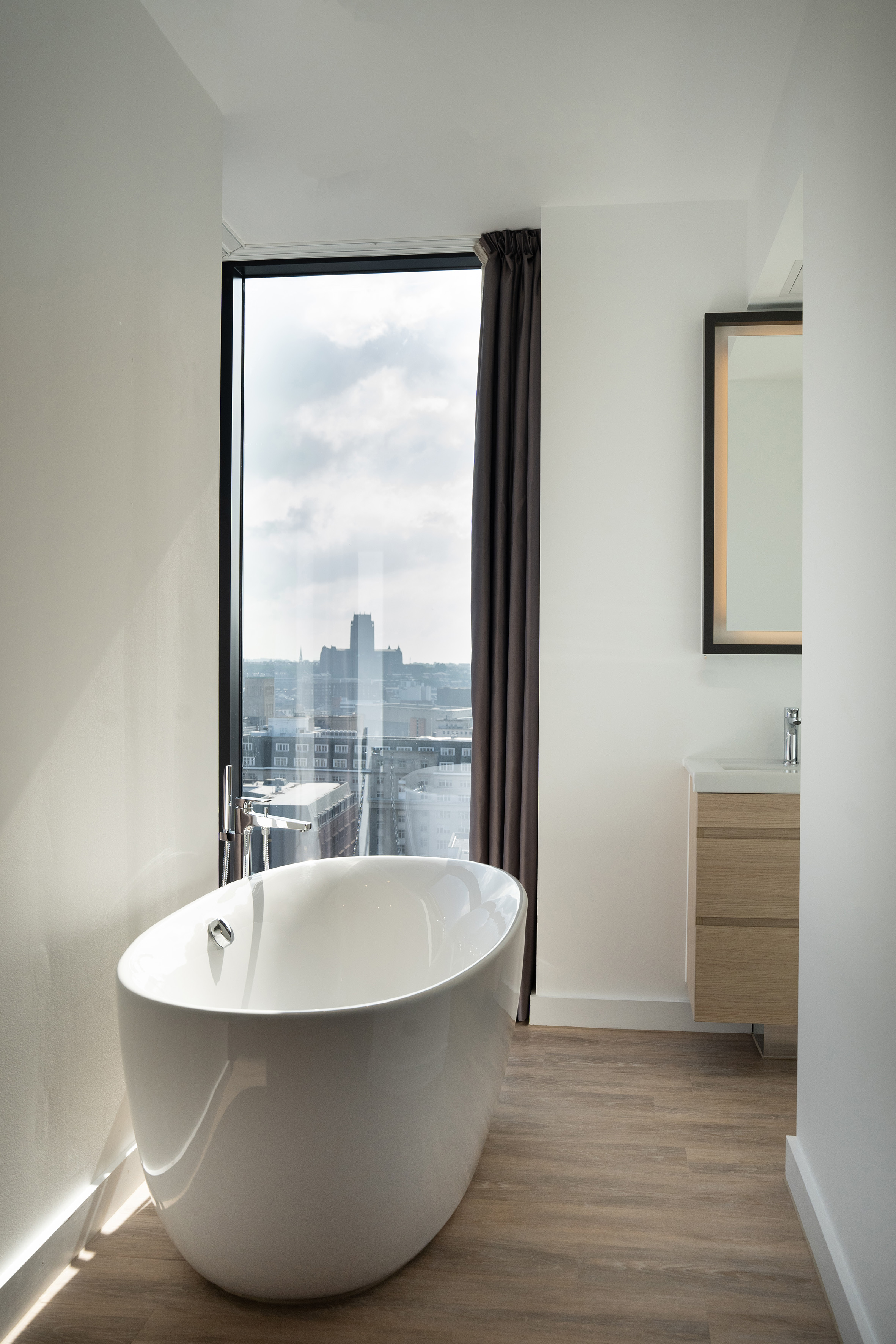 Luxury bathroom with a freestanding tub, large window city view, and wood floors.