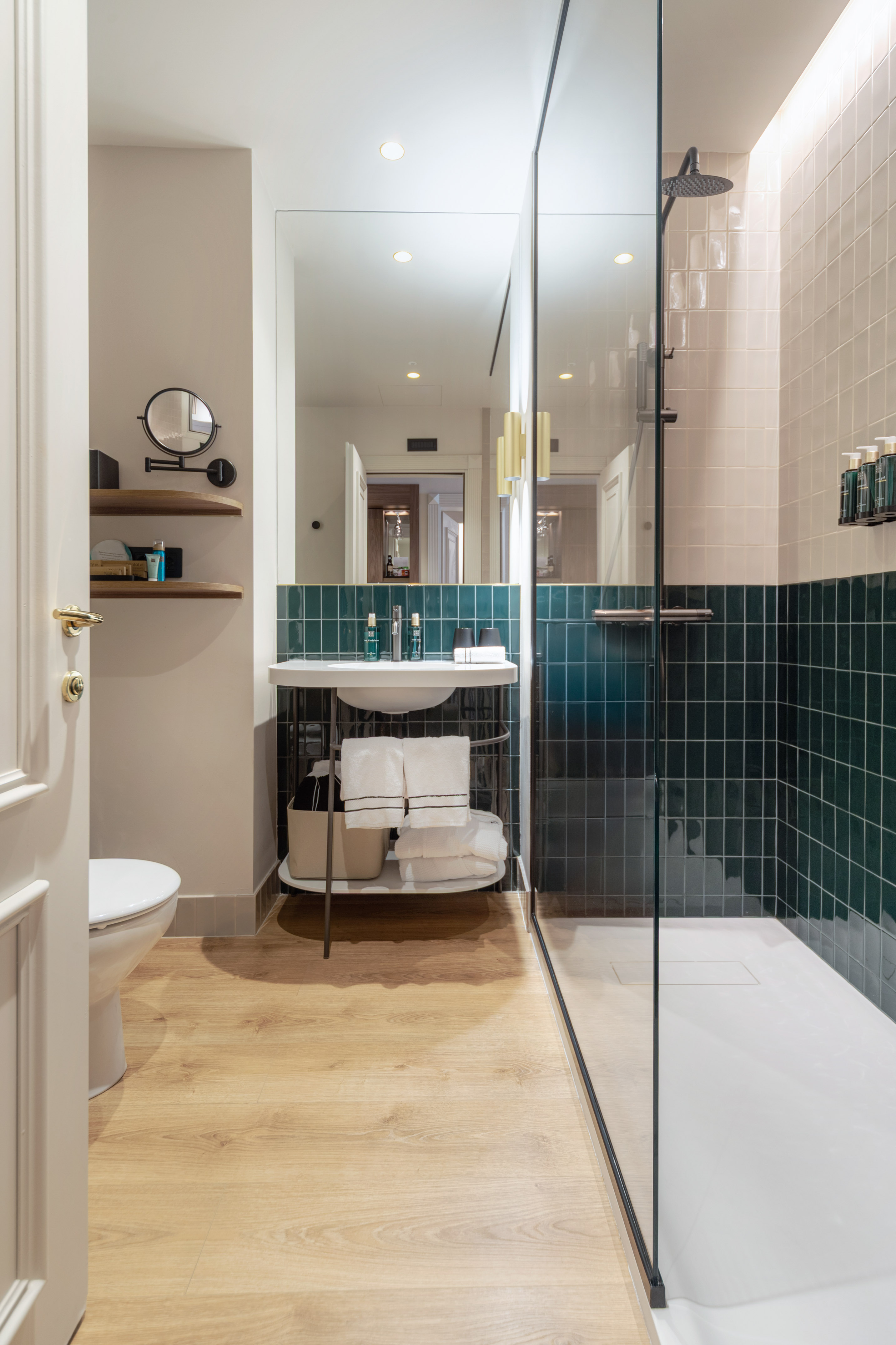Contemporary hotel bathroom with green tiled wall, glass shower, and wood floor.
