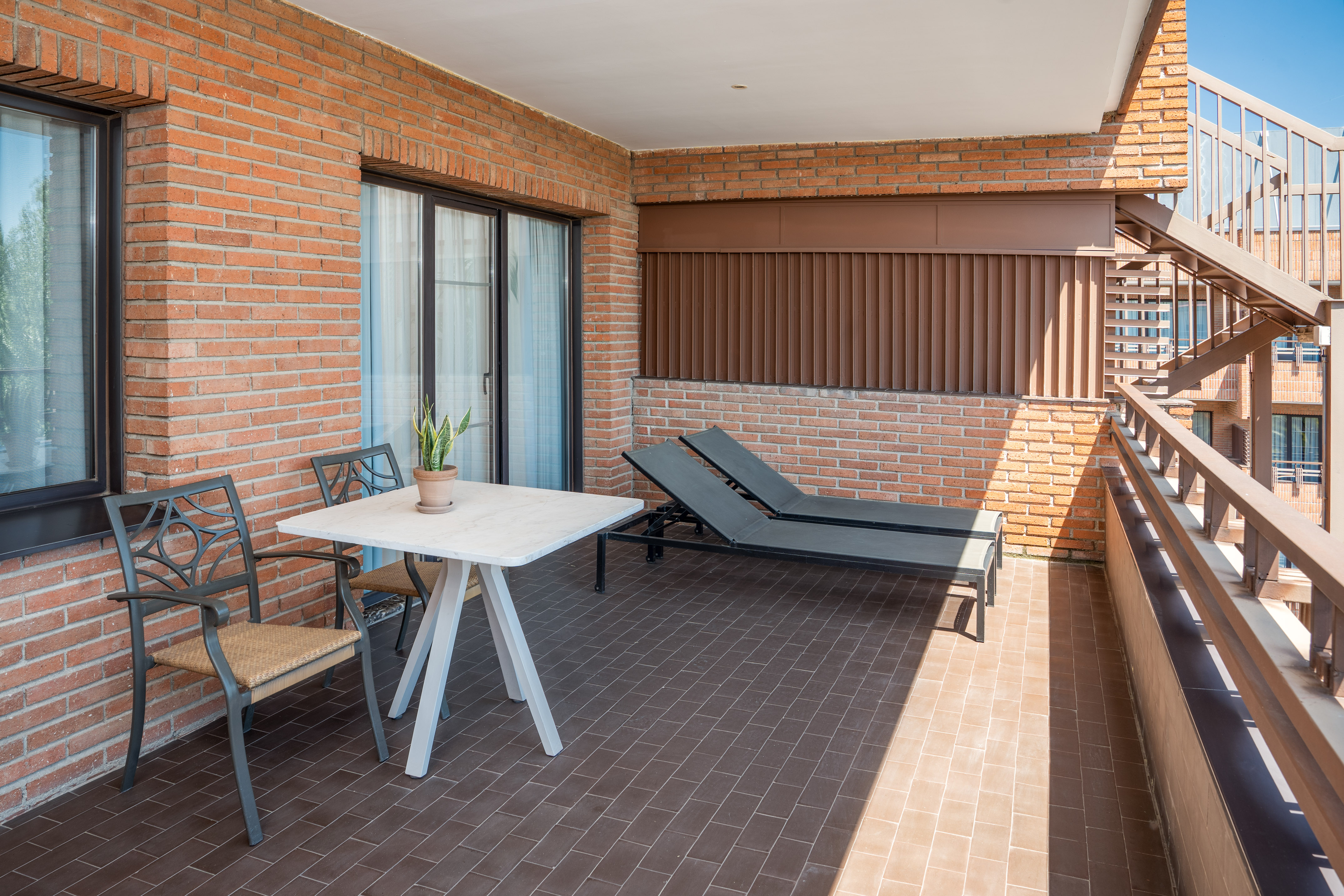 Sunny brick balcony with outdoor table, chairs, and two sun loungers on tiled floor.
