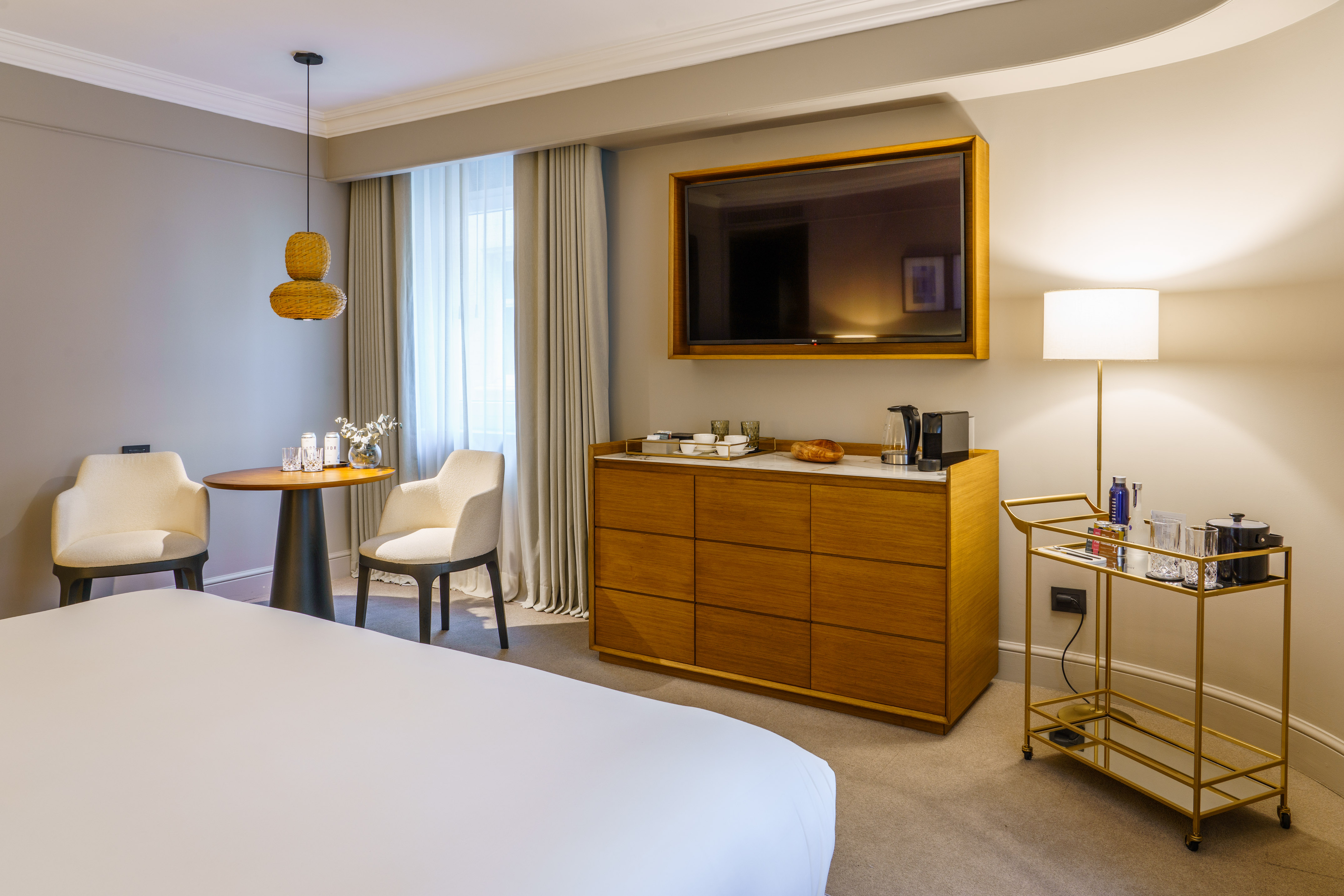 Modern hotel room interior with bed, mounted TV, coffee maker, and bar cart.