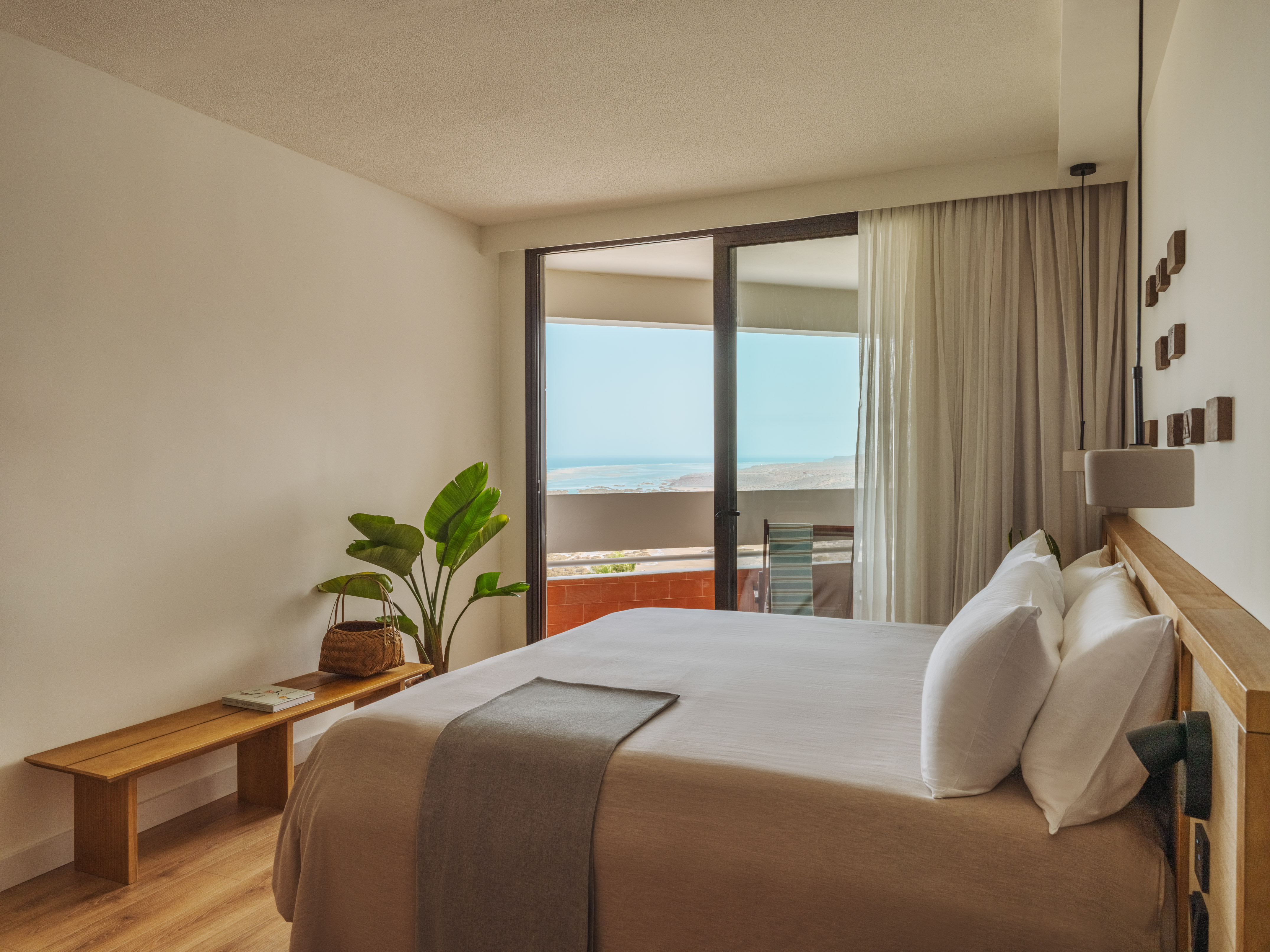 Modern coastal bedroom with ocean view balcony, plant, and wooden bench.