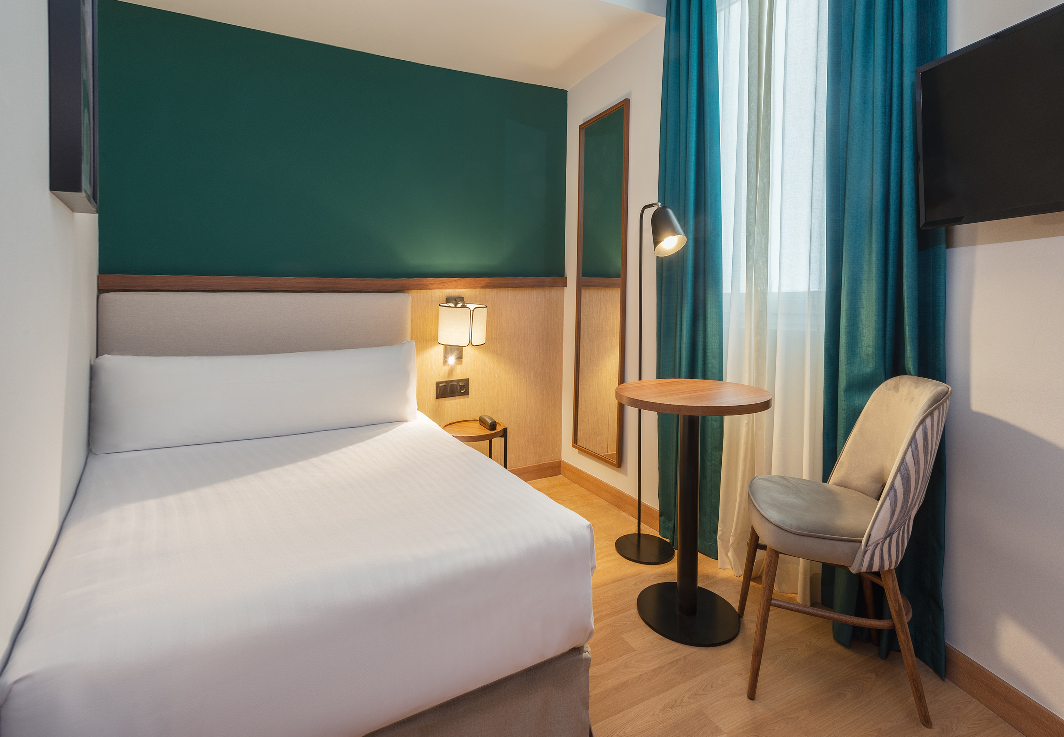 Cozy modern hotel bedroom with a single bed, dark green accent wall, and teal curtains.