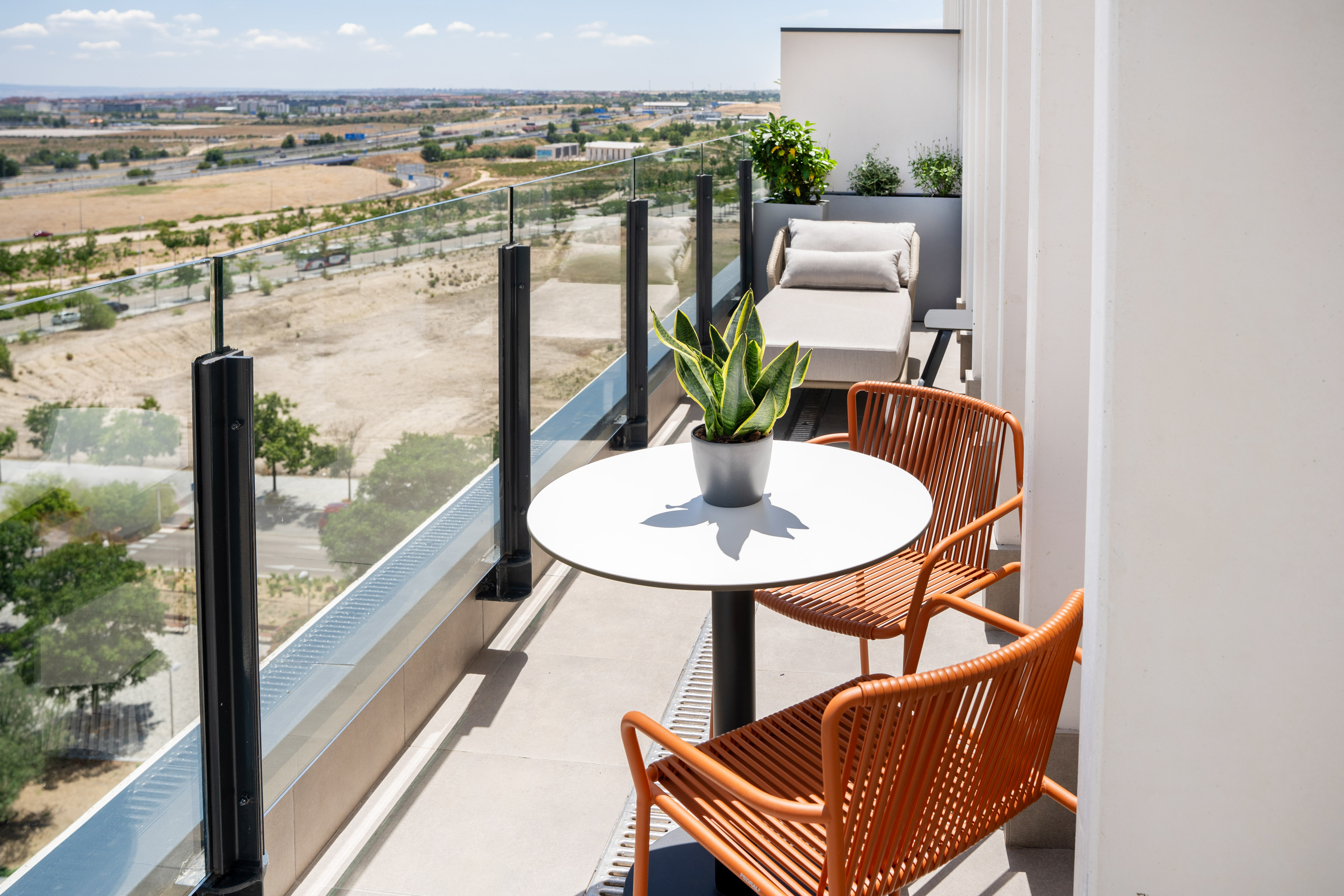 Modern balcony with city view, glass railing, orange chairs, table, and sun lounger.