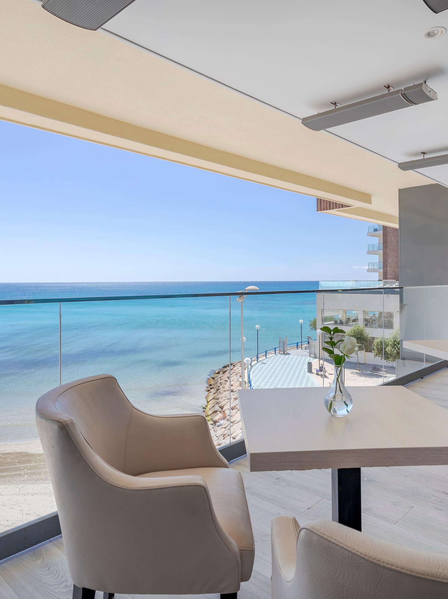 Balcón de hotel frente al mar con mesas, sillas y vistas a la playa de arena.