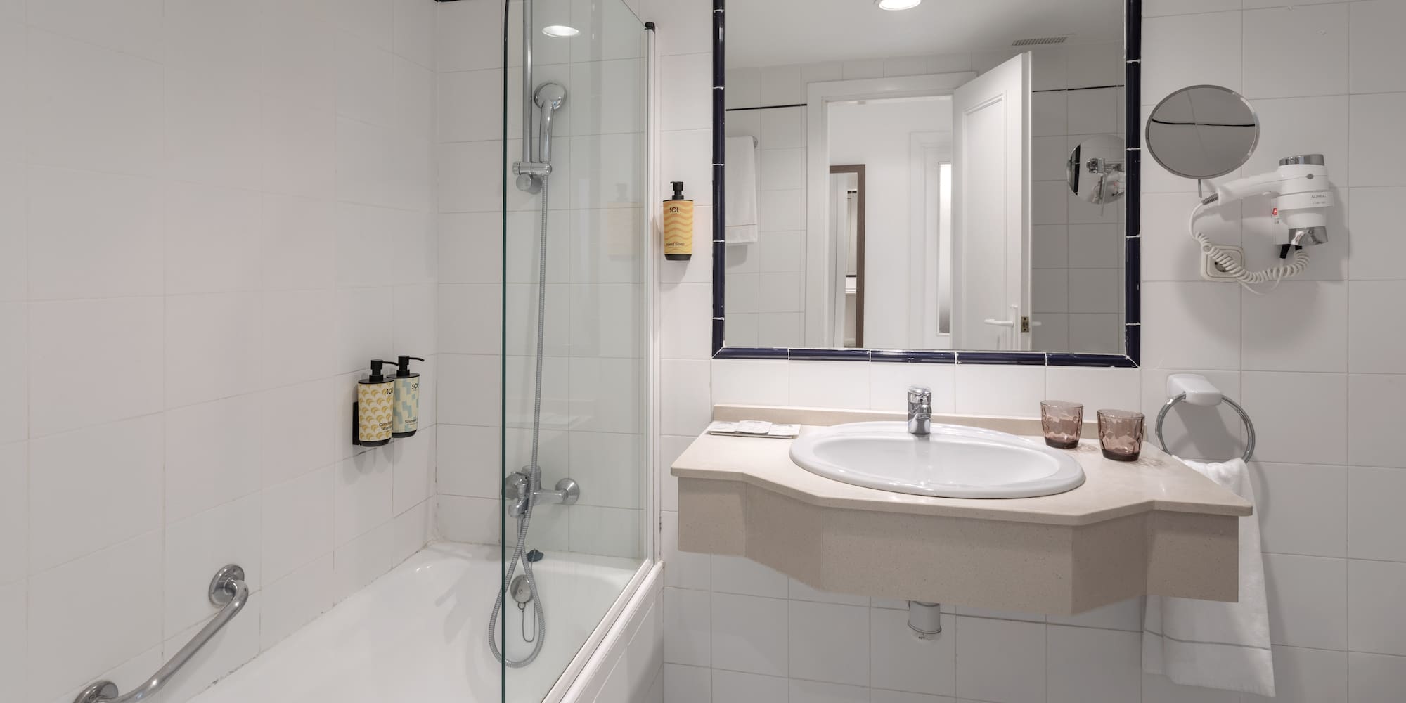 Hotel bathroom with bathtub, glass shower screen, sink, mirror, and white tiles.