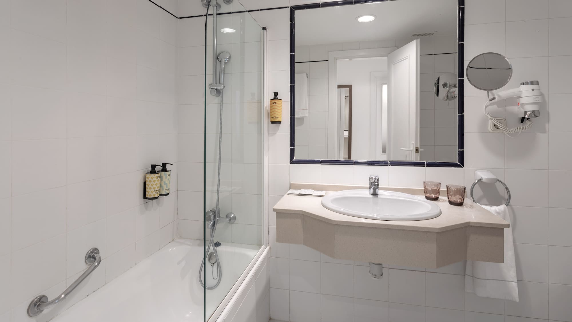 Hotel bathroom with bathtub, glass shower screen, sink, mirror, and white tiles.