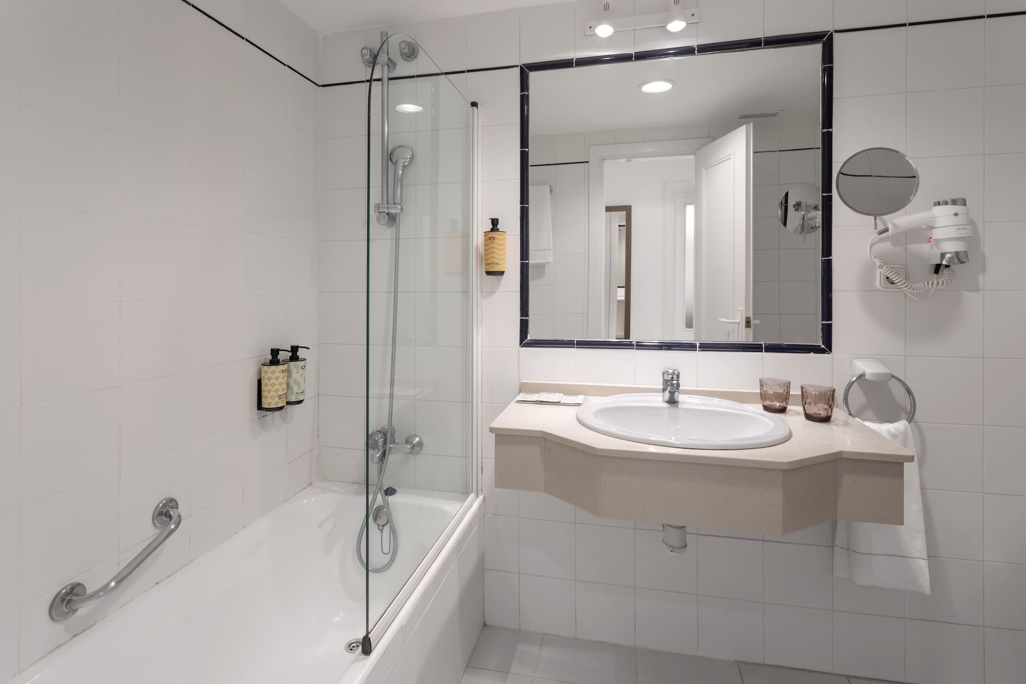 Hotel bathroom with bathtub, glass shower screen, sink, mirror, and white tiles.