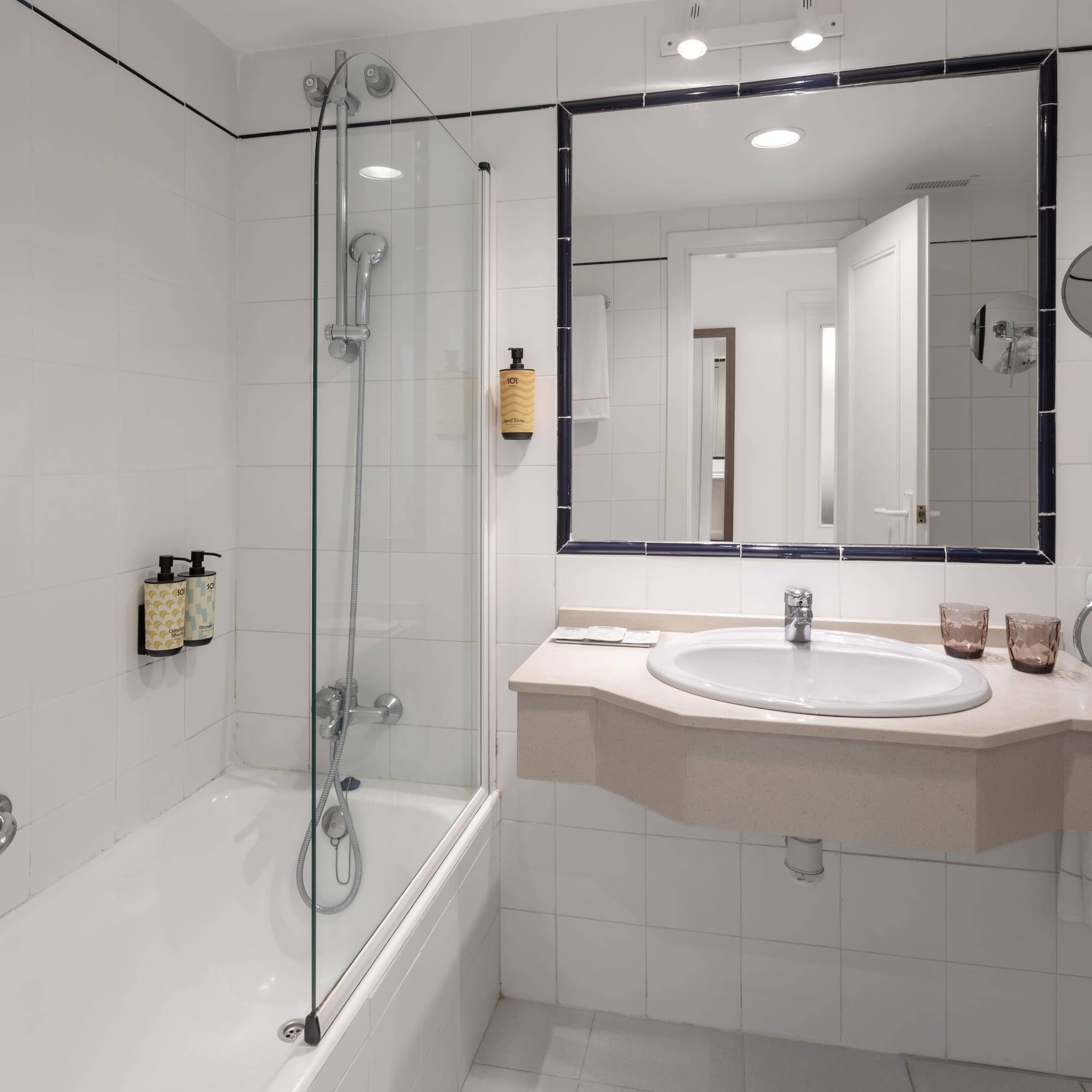 Hotel bathroom with bathtub, glass shower screen, sink, mirror, and white tiles.