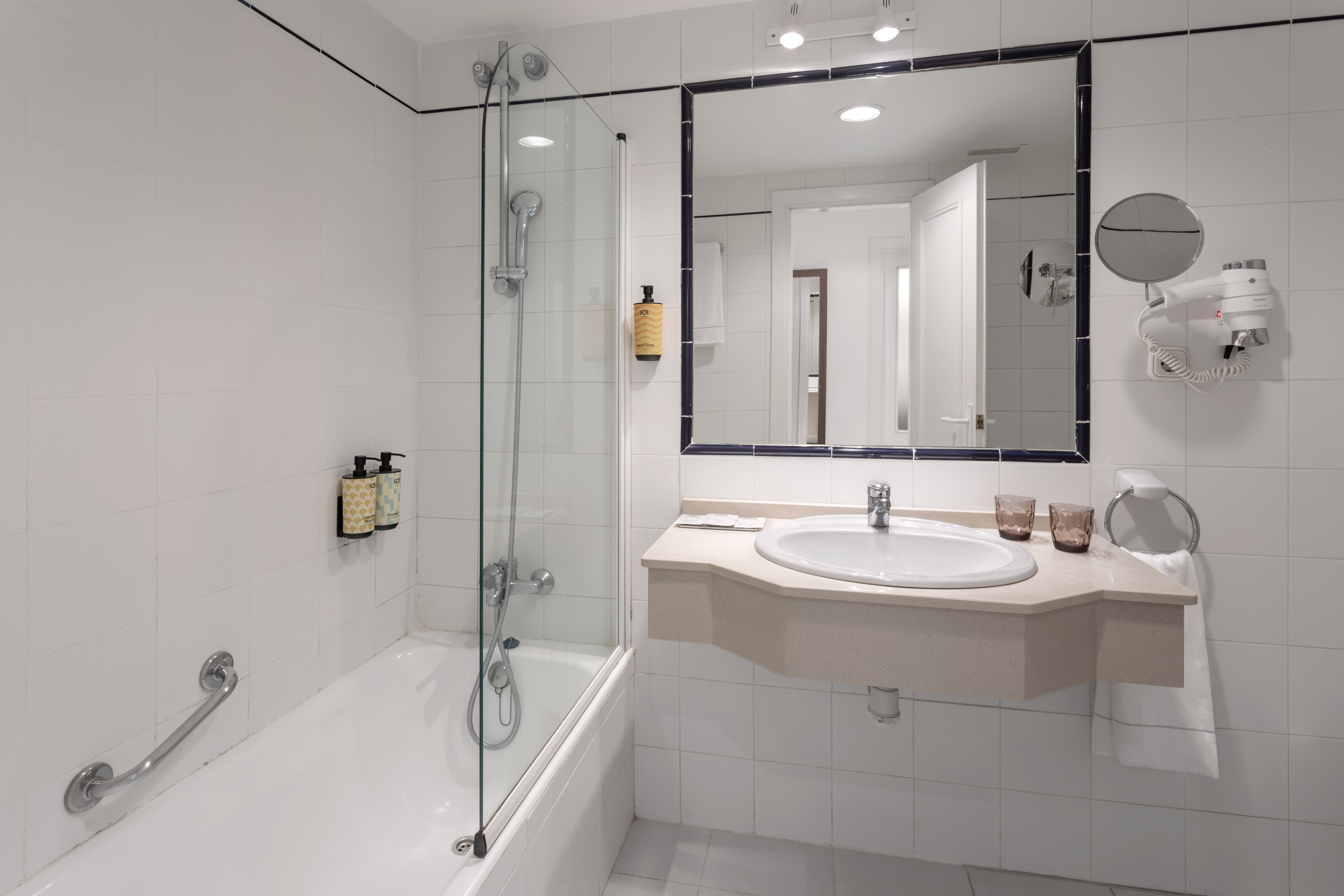 Hotel bathroom with bathtub, glass shower screen, sink, mirror, and white tiles.