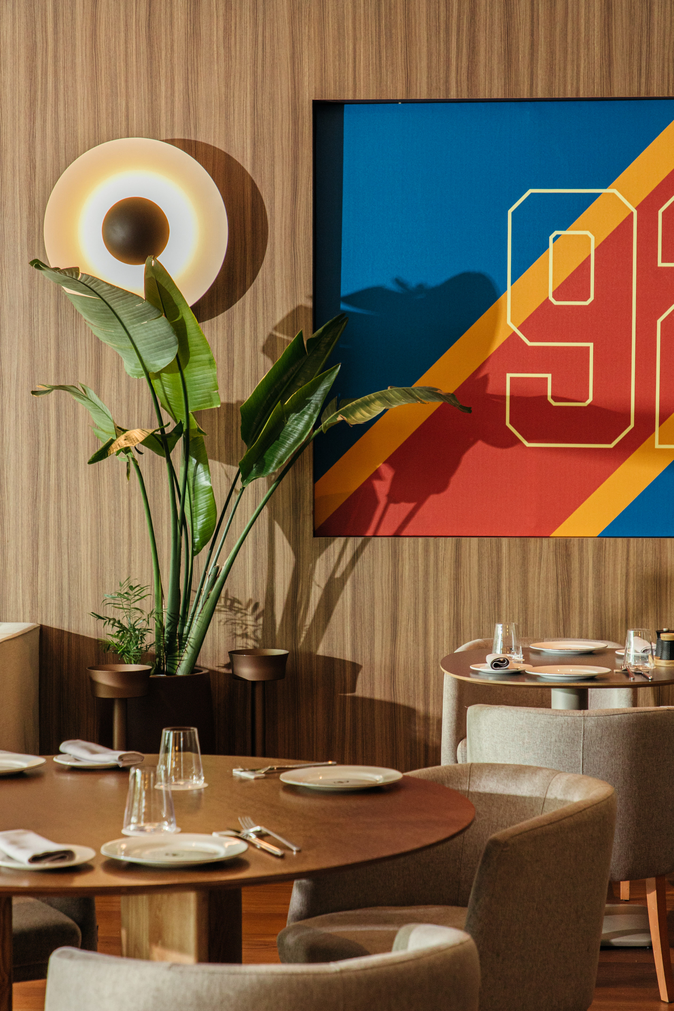 Modern restaurant with wood walls, lush plant, colorful '98' art, and dining tables.